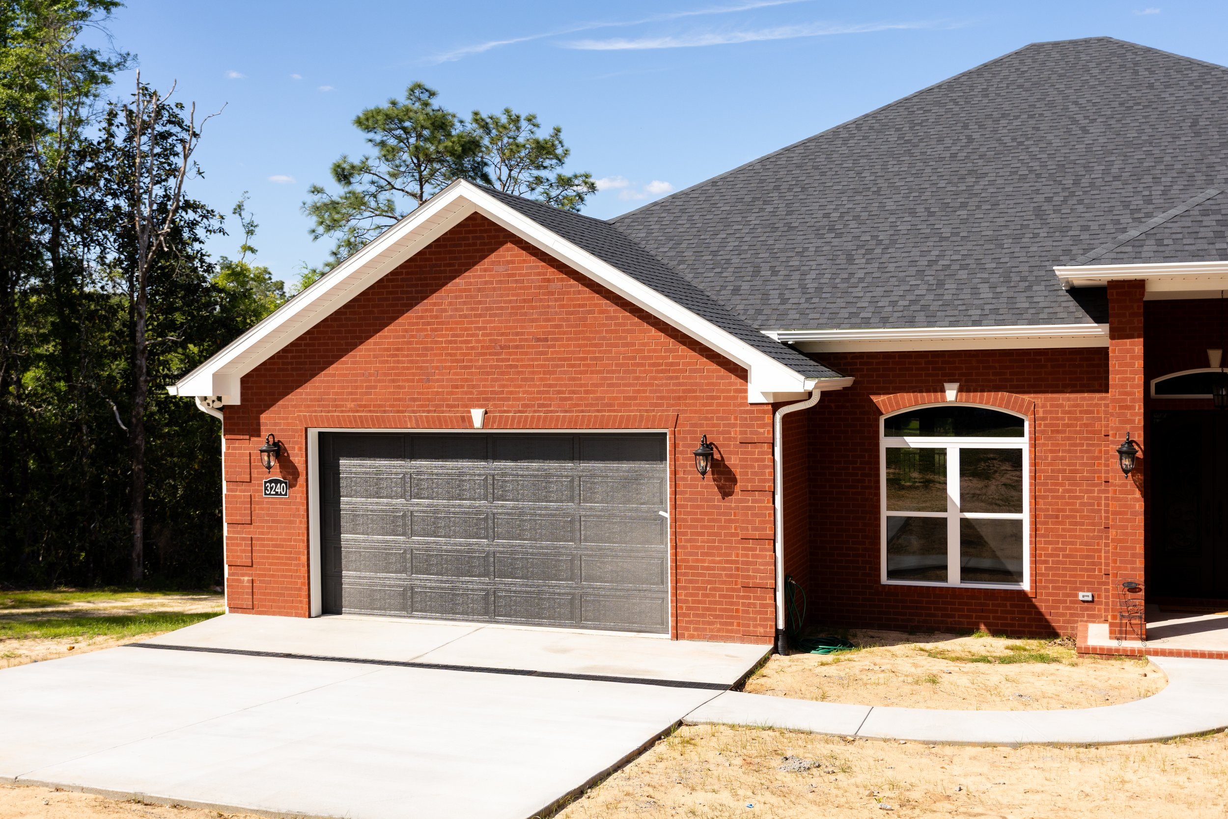 Front view of a brick house with a gray garage door, black lantern lights, and a large window, surrounded by trees, with a partly cloudy sky overhead.