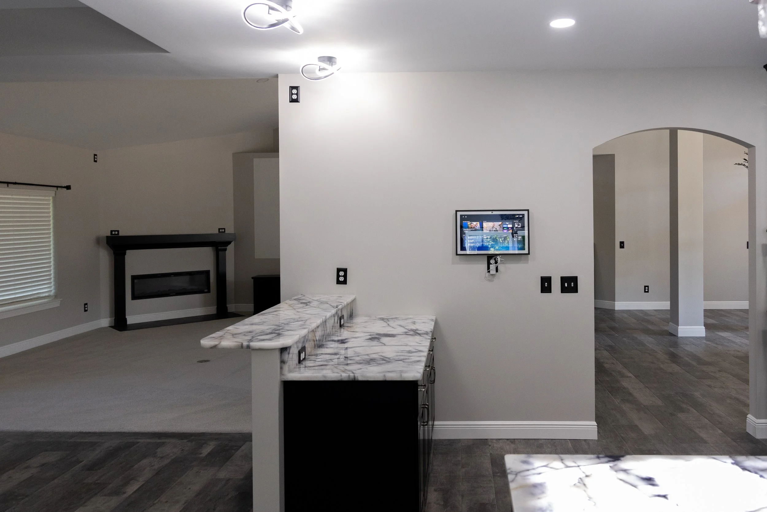 Interior view of a modern living room with a marble kitchen island, a wall-mounted TV, a fireplace, and arched doorways. The room features white walls, hardwood flooring, and ceiling lights.