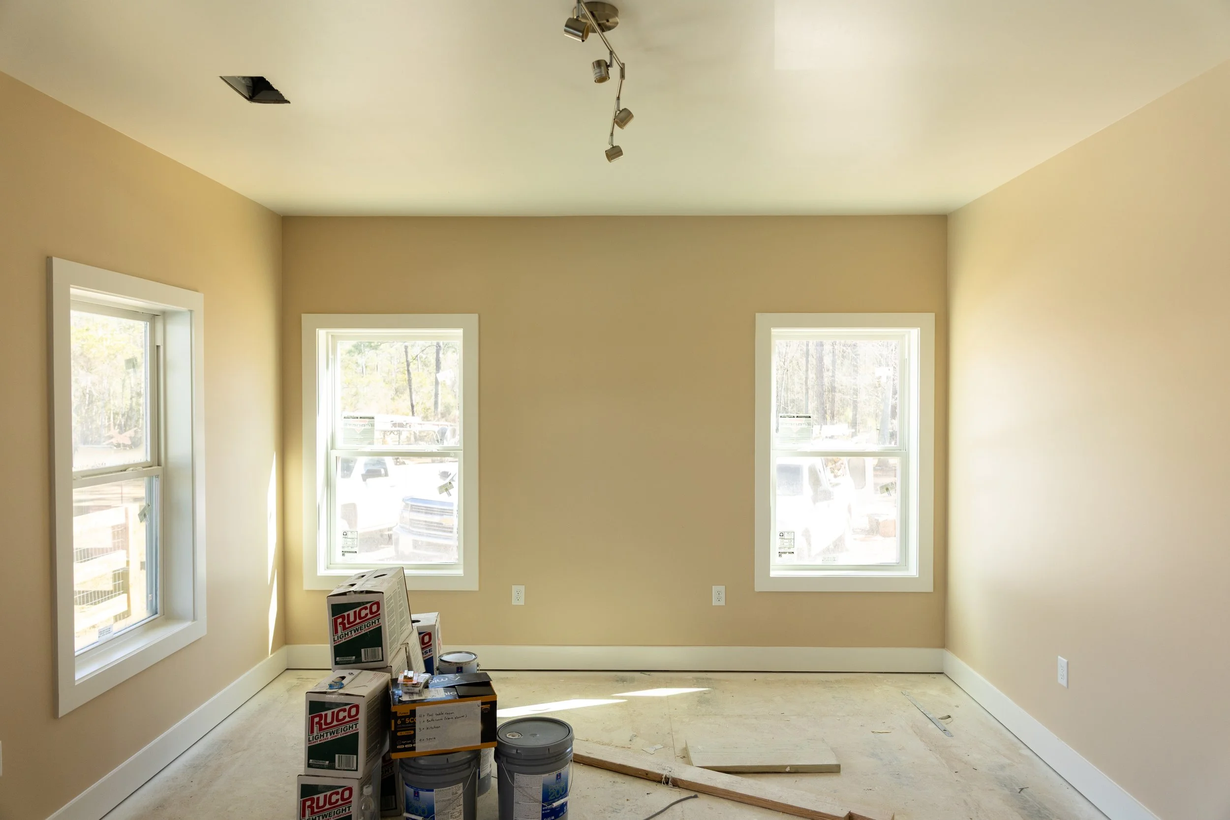A room under construction with beige walls and three large windows. Construction boxes and materials are on the floor.