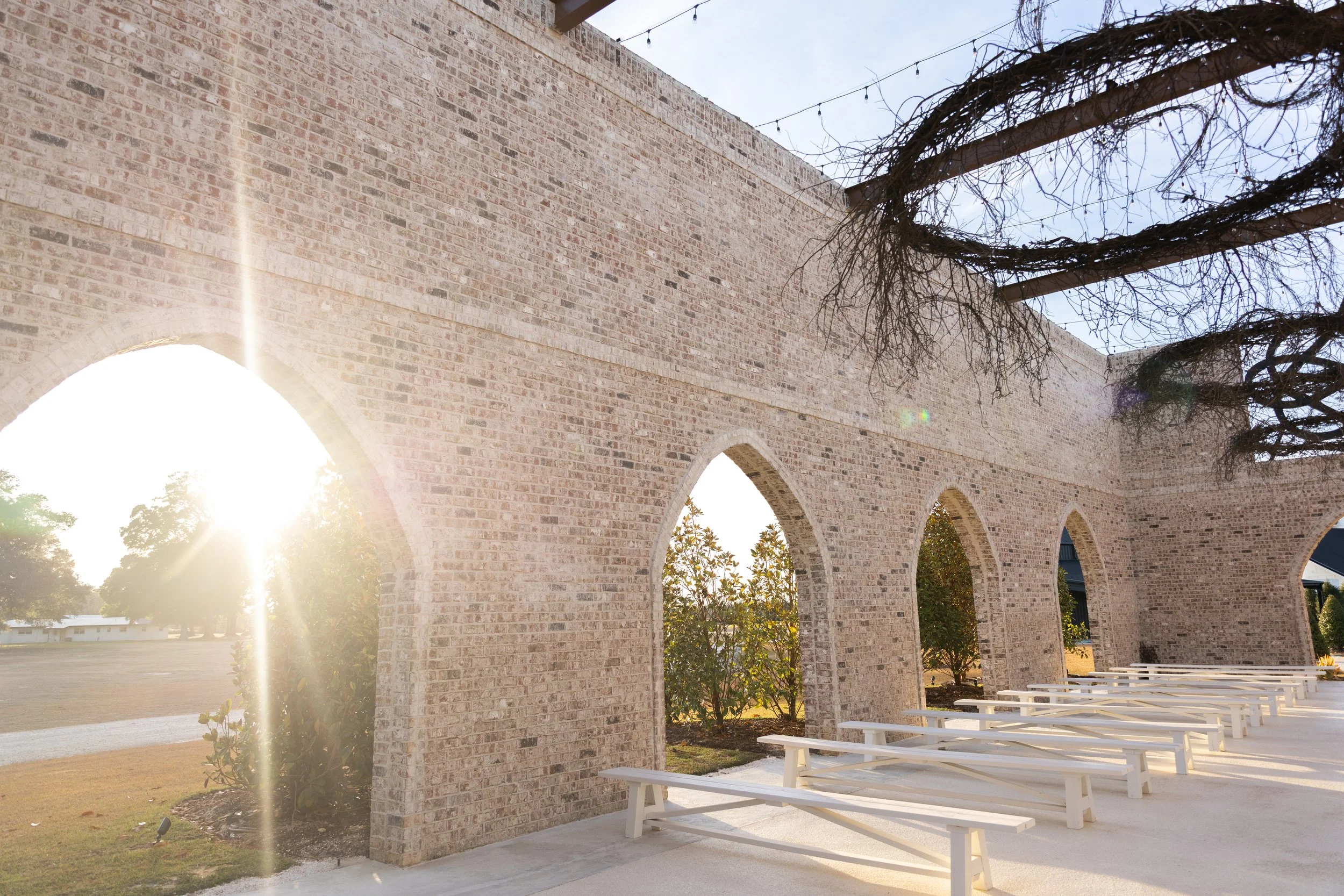 Sunset light shines through a row of brick arches in an outdoor seating area with white benches and trees with sparse branches.