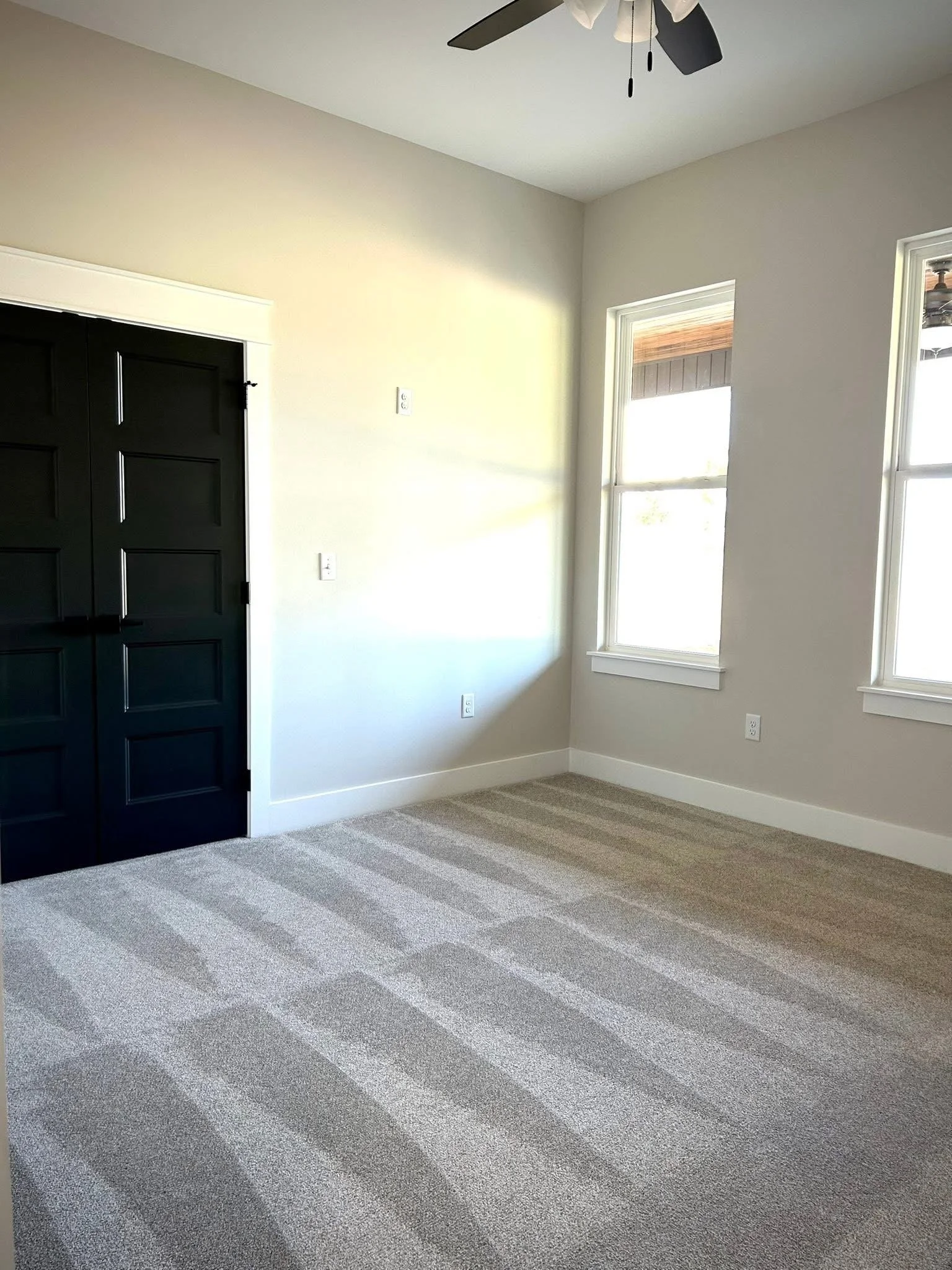 Empty room with beige walls, brown striped carpet, two windows, a black ceiling fan, and white door trim.