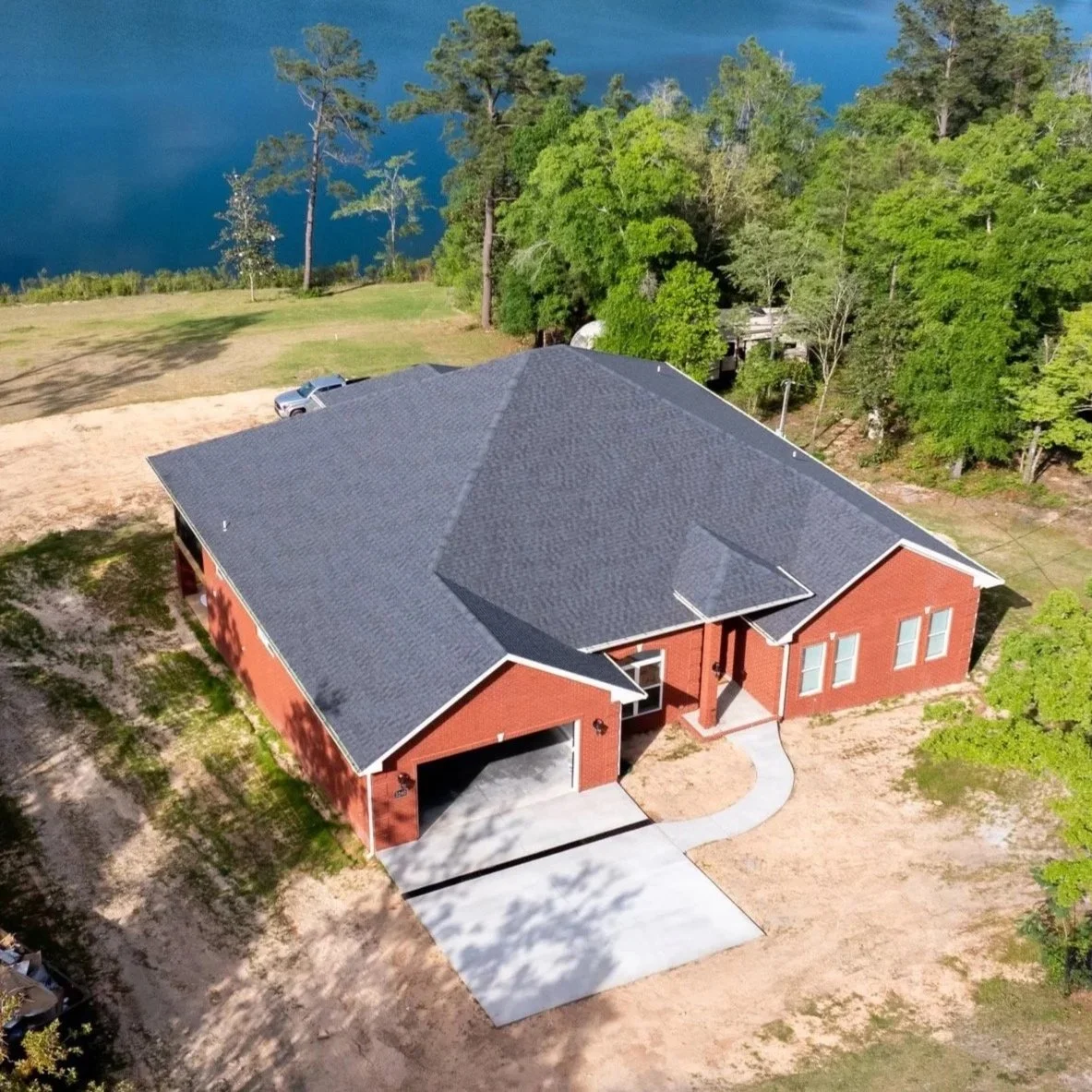 Aerial view of a single-family brick house with a dark gray pitched roof, in a green, wooded area near a body of water.