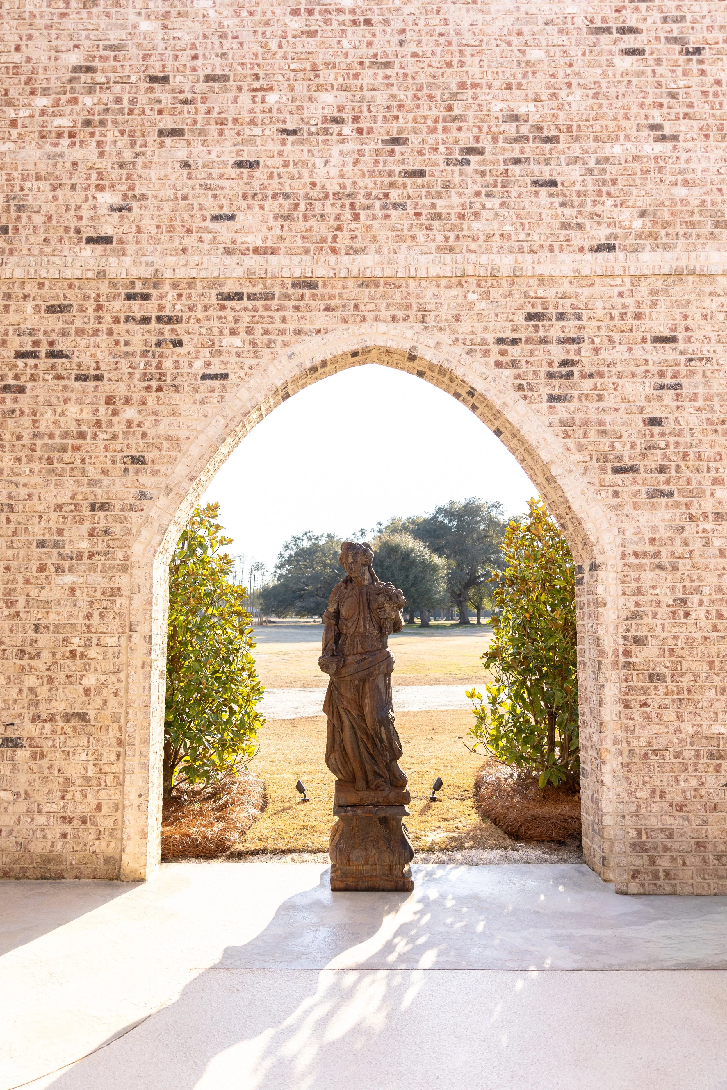 A view of a brick archway with a wooden statue of a woman in a long dress, standing on a pedestal, framed by greenery, with trees and a grassy field in the background, sunlight casting shadows.