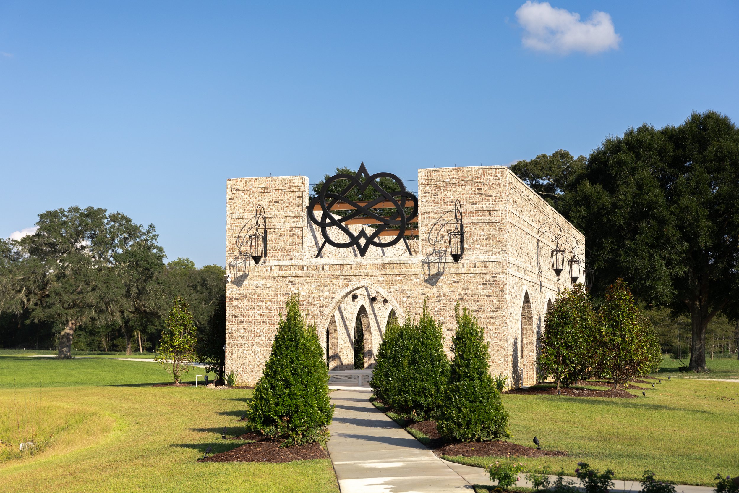 A stone building with an ornate metal design on top, surrounded by manicured bushes and a paved walkway leading to the entrance, with trees and a blue sky with clouds in the background.