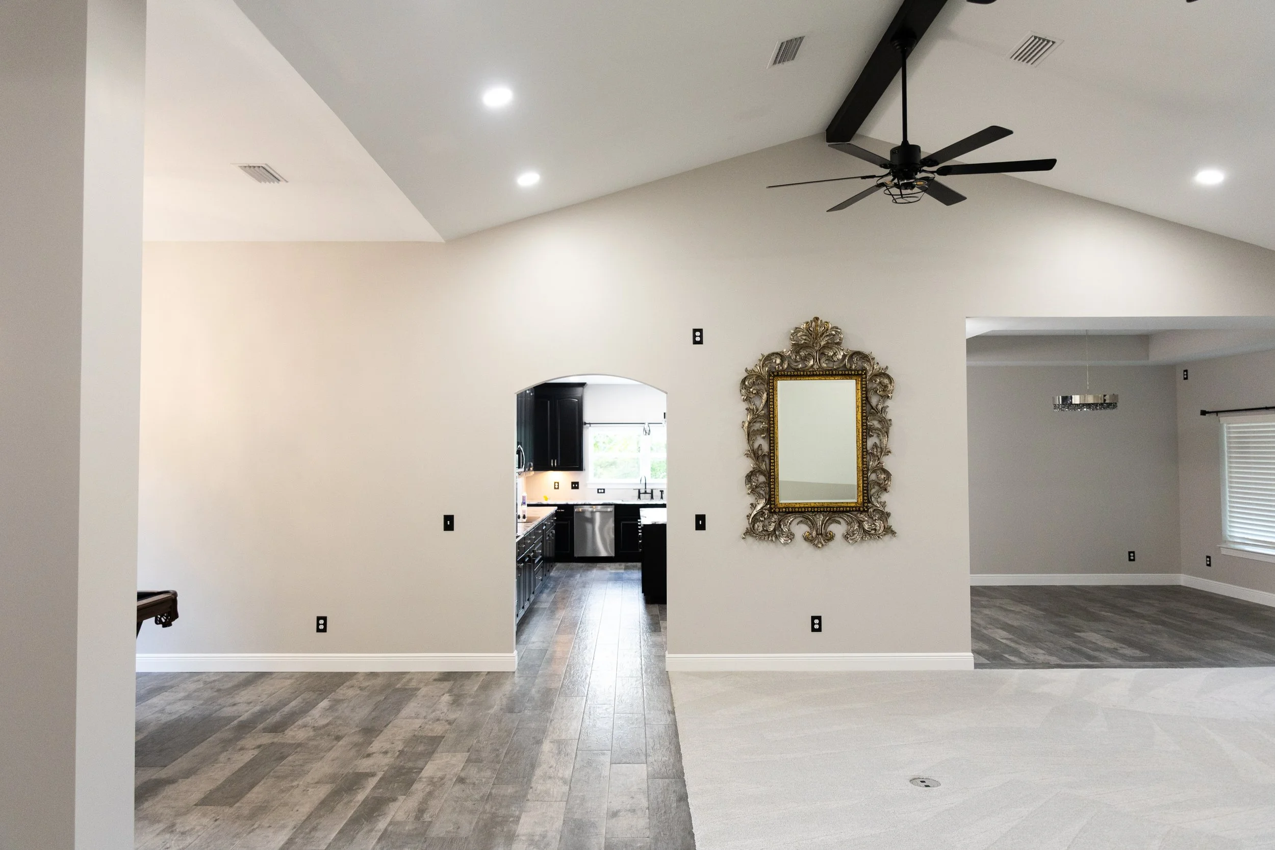 Open-concept living area with a white ceiling, black ceiling fan, and a large ornate gold-framed mirror on a light beige wall. The living area flows into a kitchen visible through an arched doorway, with black cabinets and a window. The flooring vari