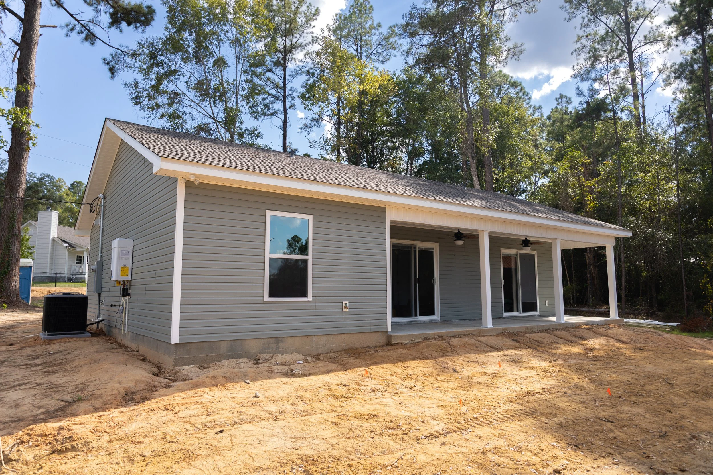 Newly constructed house with grey siding, sliding glass doors, and a covered patio, surrounded by trees and a dirt ground.
