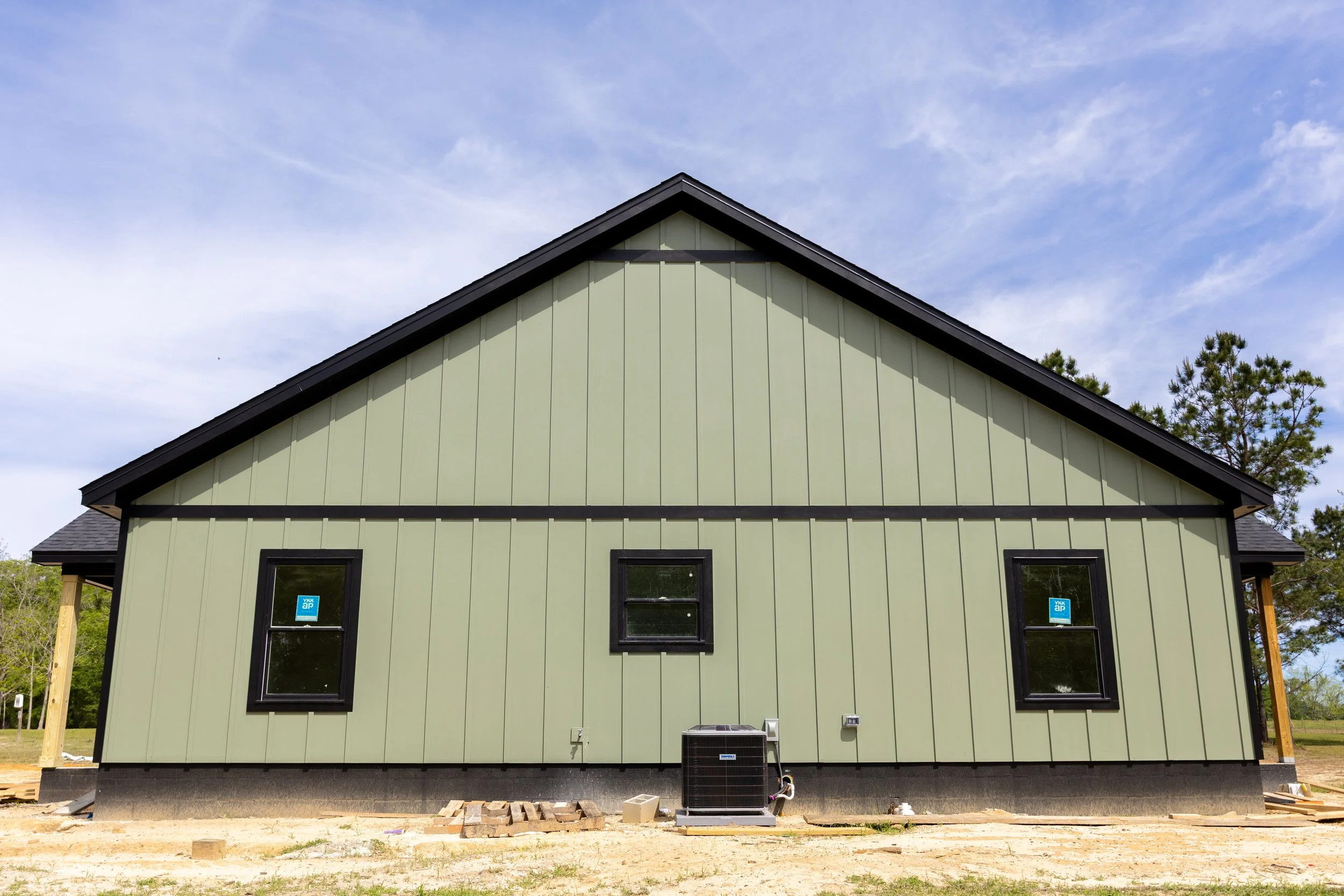 A two-story house under construction, painted light green with black trim, has several black-framed windows and is situated outdoors on a partially constructed foundation with building materials and an HVAC unit in front. The background features a bl