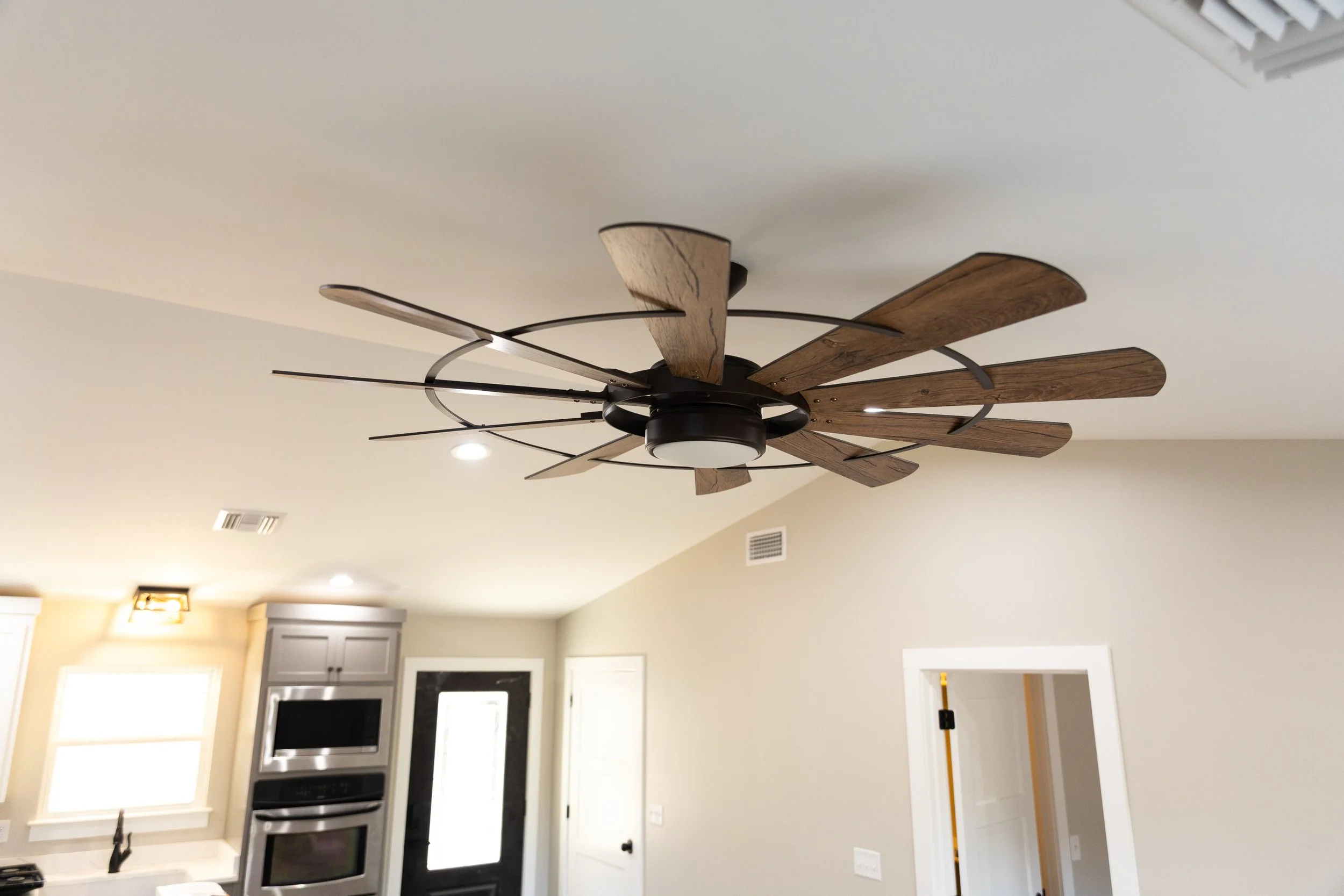 Ceiling fan with wooden blades and integrated lights in a modern kitchen, ceiling and wall with windows, kitchen appliances, and cabinets visible.