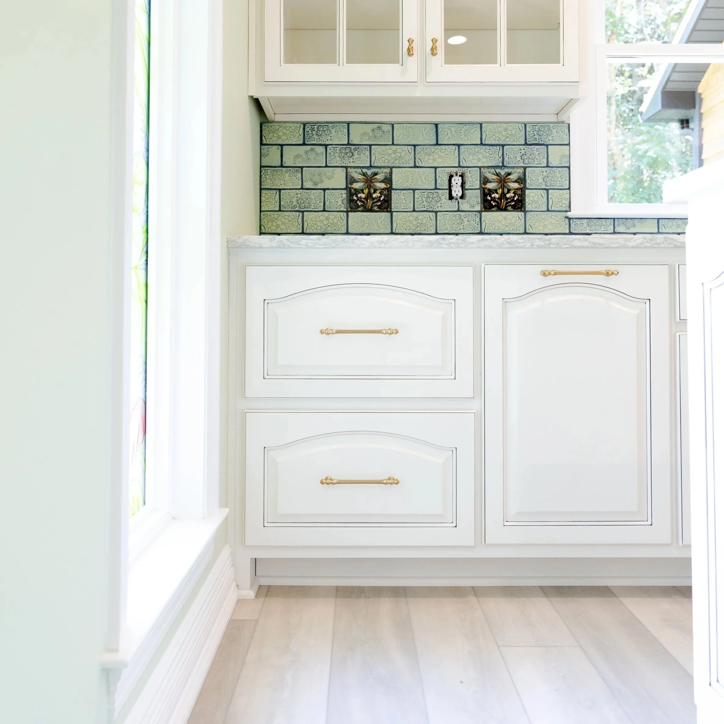 A kitchen with white cabinets, a tiled backsplash with floral and dragonfly designs, a window with greenery outside, and light-colored wooden flooring.