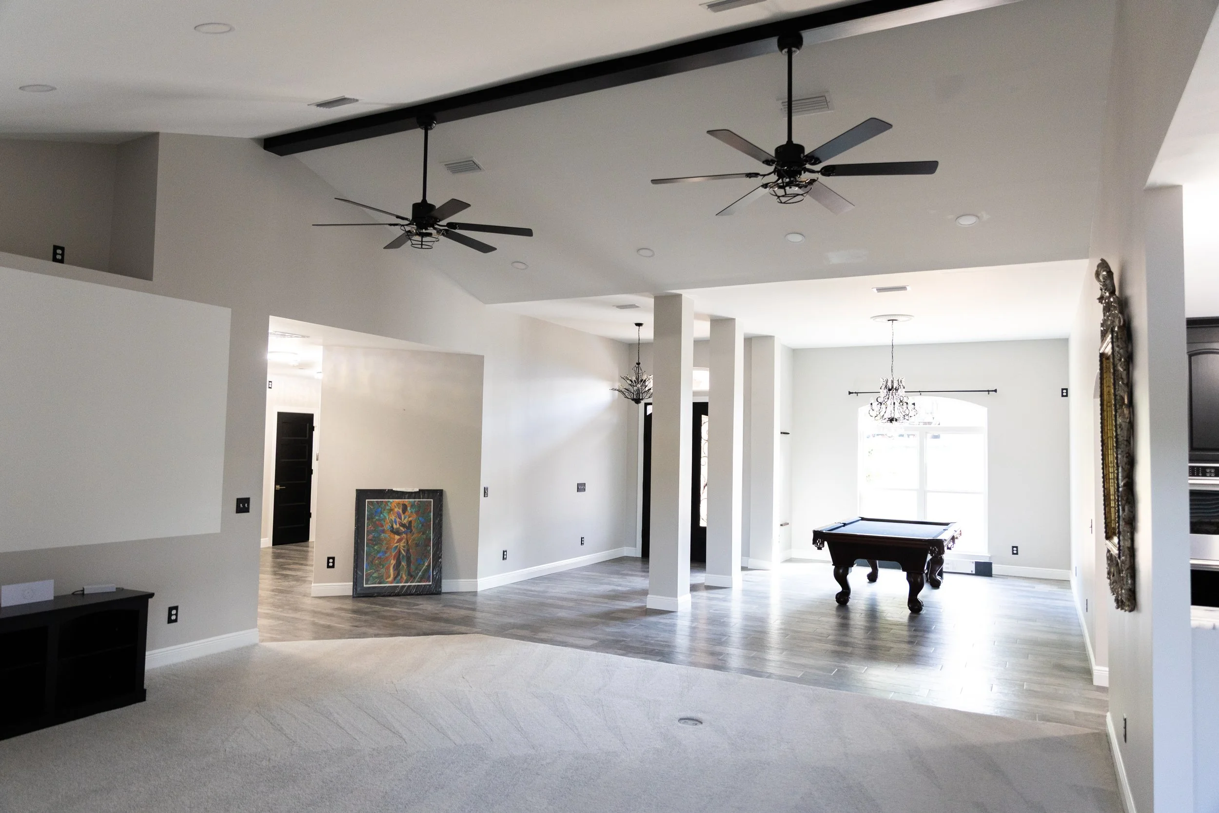 Interior view of a spacious modern house with high ceilings, ceiling fans, a chandelier, and a pool table, with a mix of carpeted and hardwood floors, and artwork leaning against the wall.