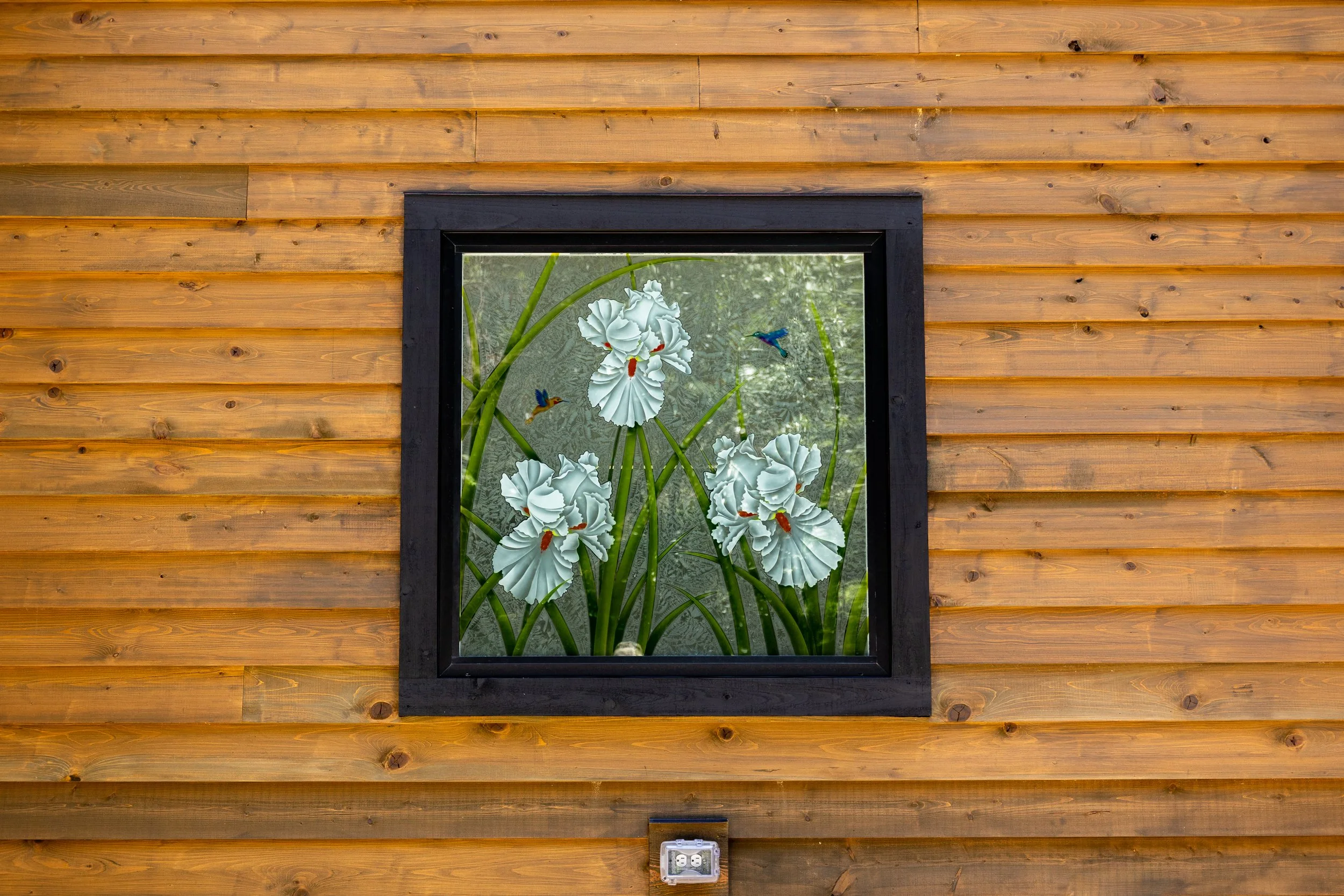 A window with decorative stickers of white irises and hummingbirds, set in a black frame on a wooden wall.
