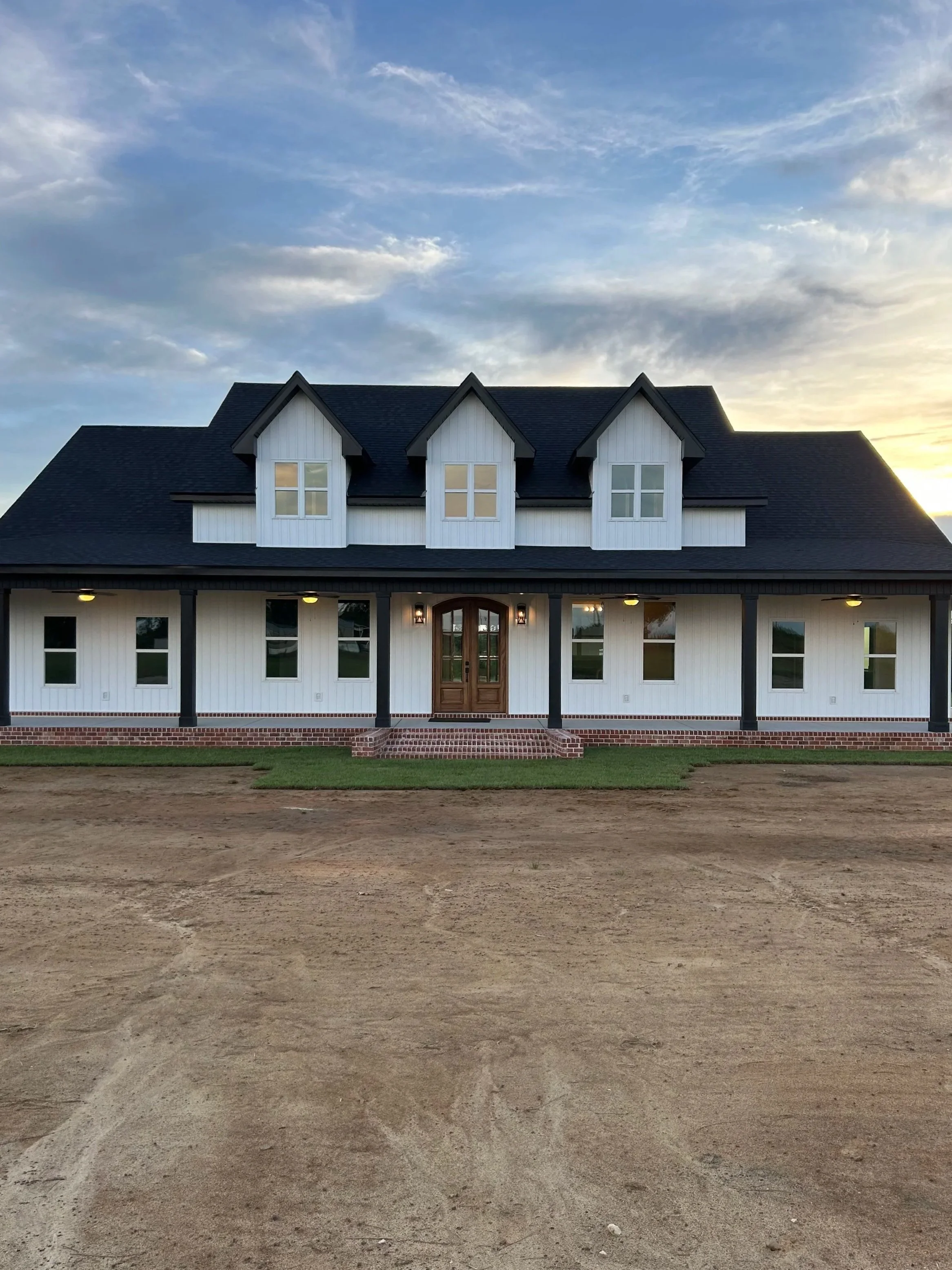 A large two-story house with a black roof, white walls, and three dormer windows. The house has a front porch with black columns, a wooden front door, and multiple windows. The ground in front is bare dirt with no landscaping.
