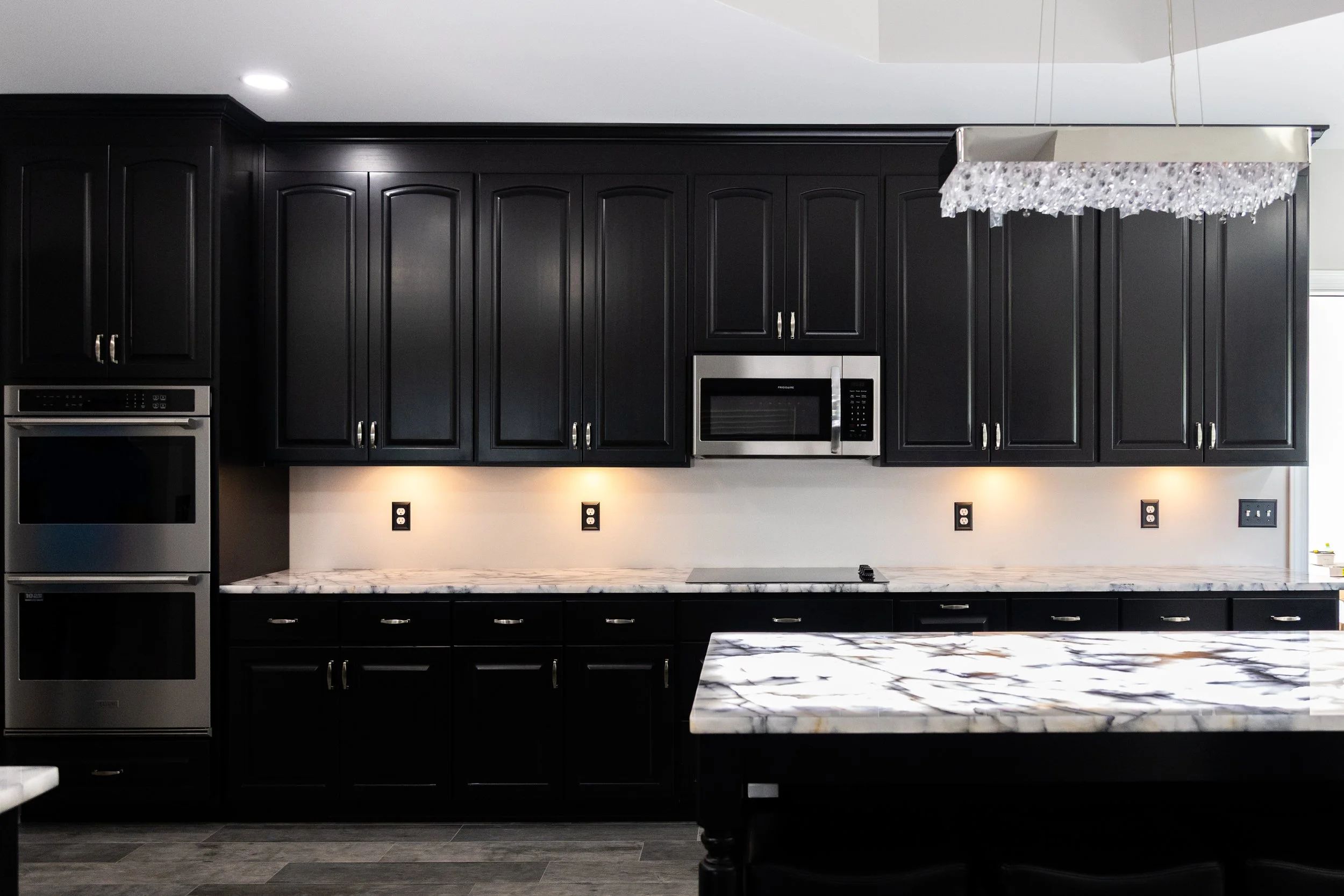 Modern kitchen with black cabinets, a marble countertop, and stainless steel appliances including a microwave and double oven. Overhead lighting and a decorative chandelier are also visible.