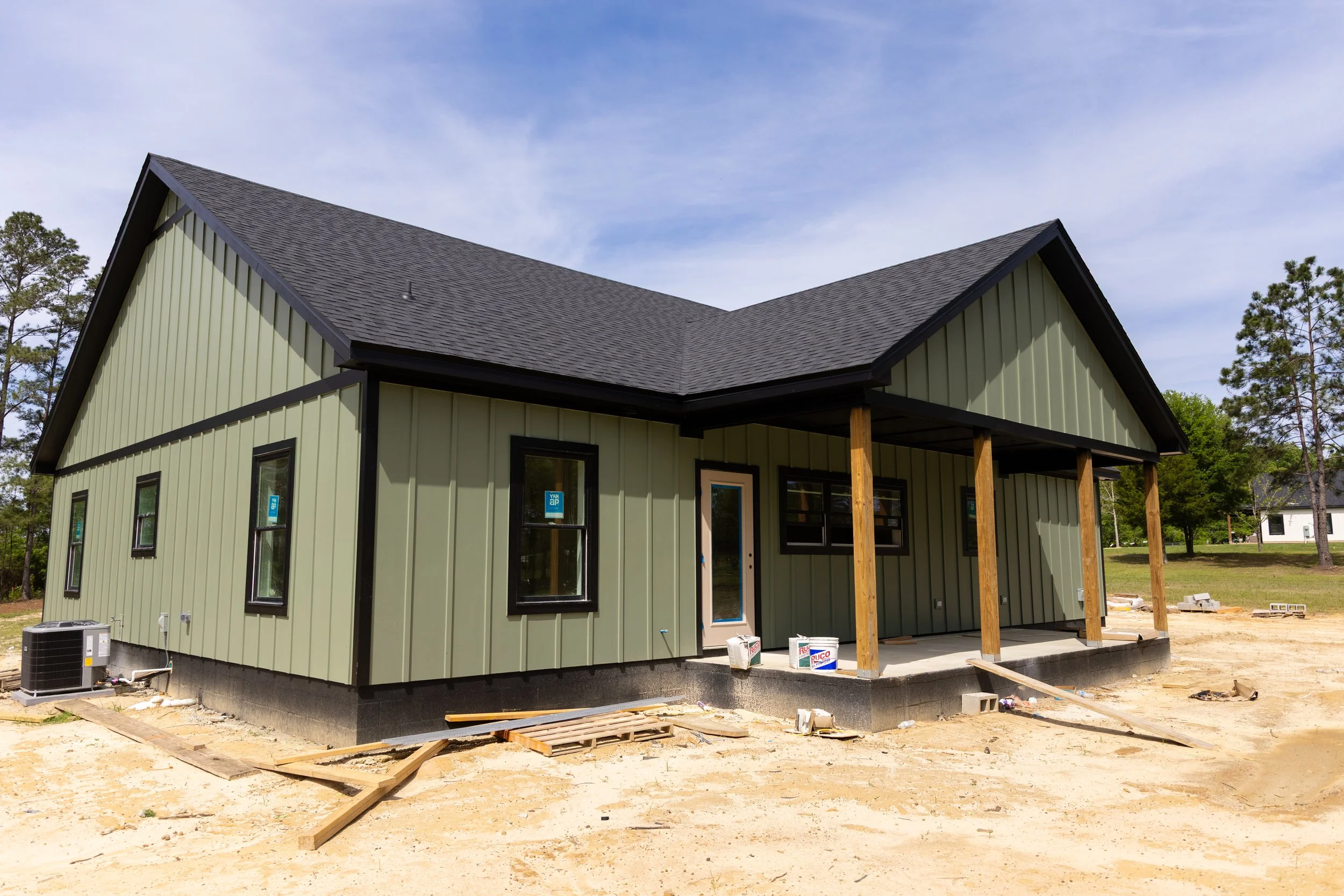 A house under construction with light green exterior walls, a gray pitched roof, and a small porch supported by wooden posts. Construction materials and tools are on the ground, and trees are in the background under a partly cloudy sky.