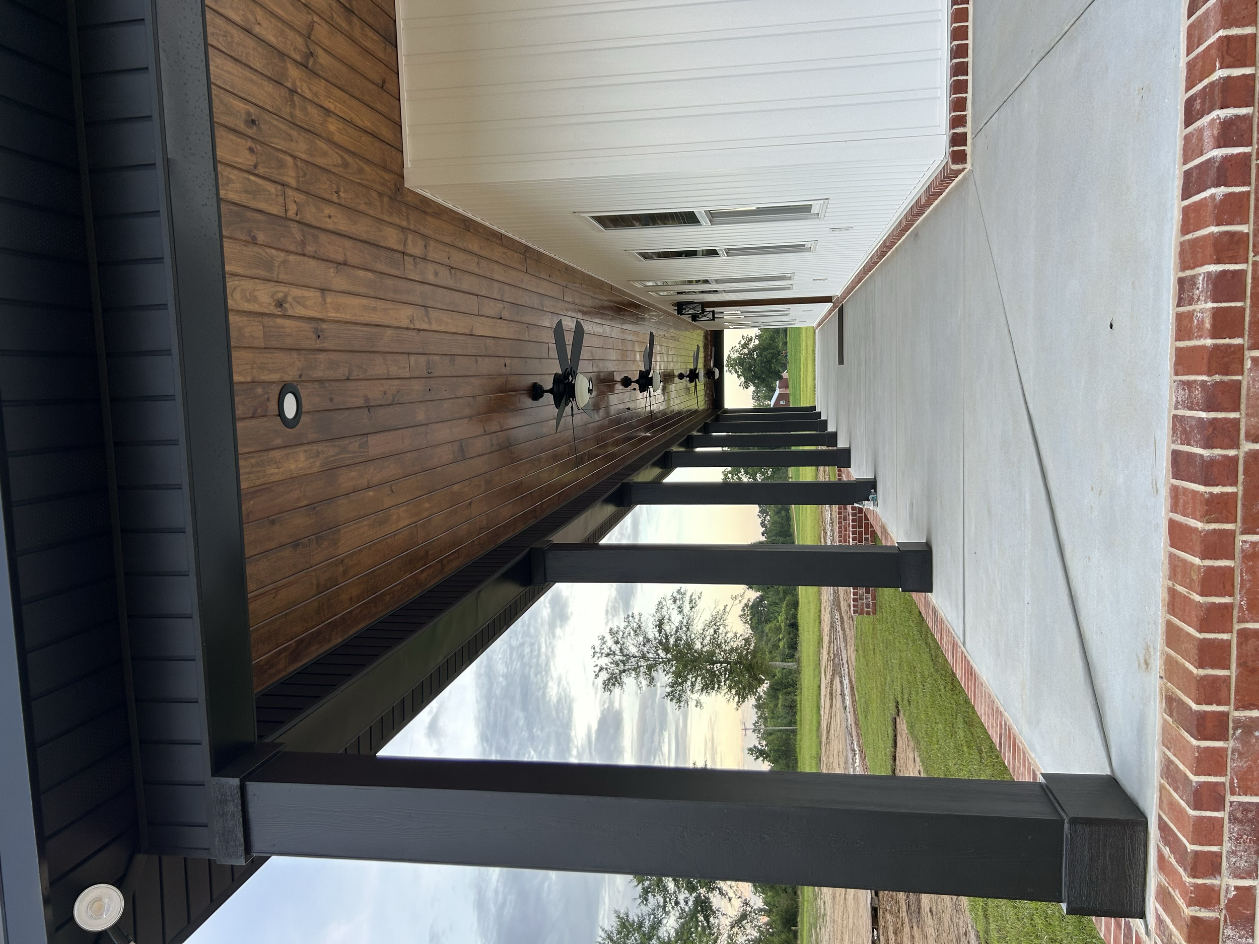 A porch area with wooden flooring on one side and concrete pavement on the other, bordered by a brick edge. The porch is partially covered with a metal and wood roof, and has black support beams. There are multiple small rectangular windows on the wh