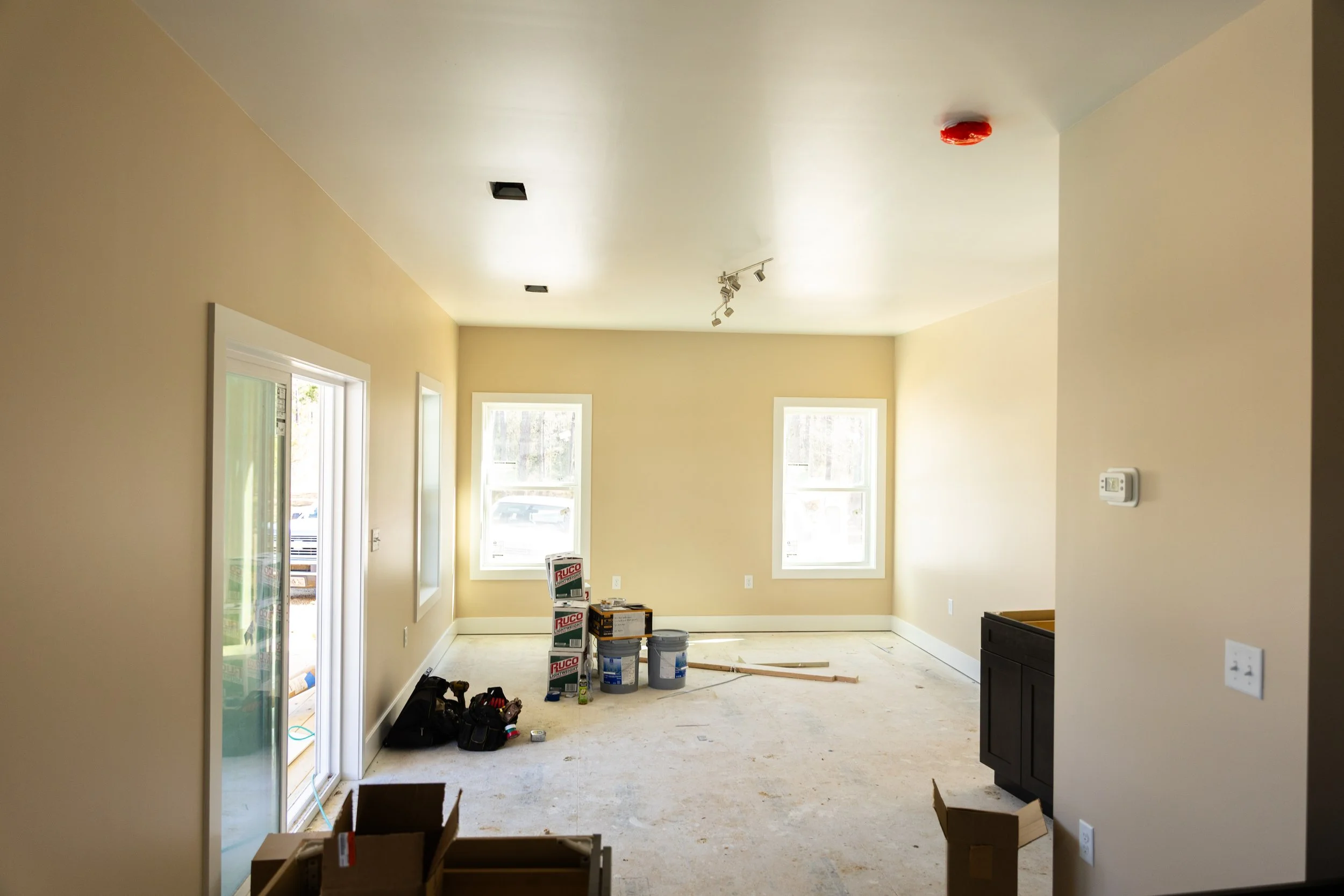 Interior room under construction with bare floors, uninstalled fixtures, and boxes of supplies, with sunlight coming through windows and a sliding glass door.