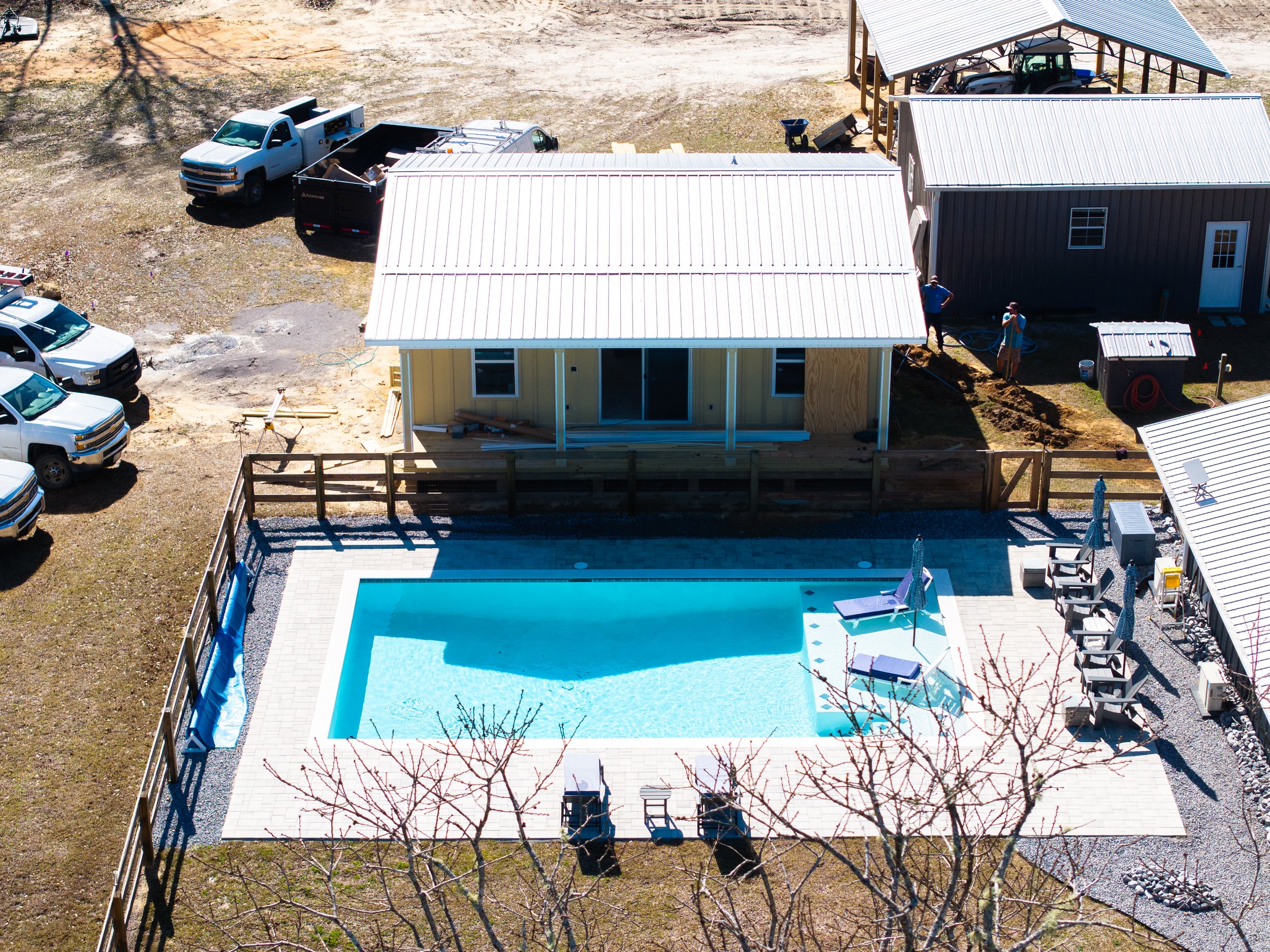 A backyard scene showing a swimming pool with a tiled deck, two lounge chairs with umbrellas, and a backyard fence. There are three houses under construction with metal roofs, construction workers, trucks, and construction materials. The yard is fenc