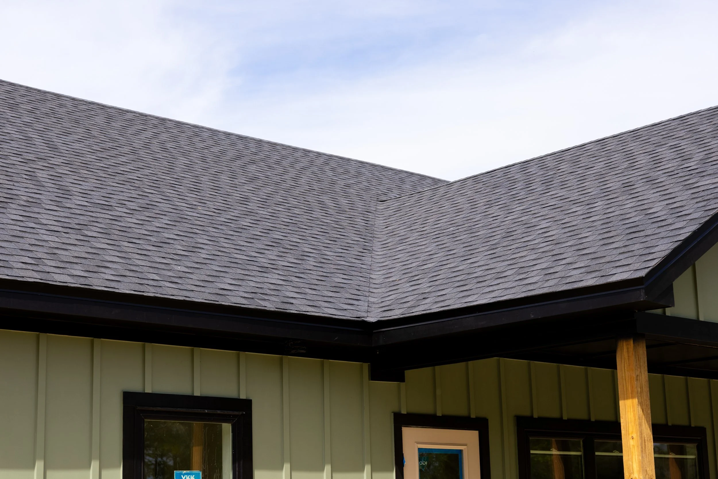 The image shows a house corner with a roof covered in gray shingles and a part of the house walls painted in light yellow-green. There are two windows visible, with black frames. One window is larger and the other smaller, with the larger window havi