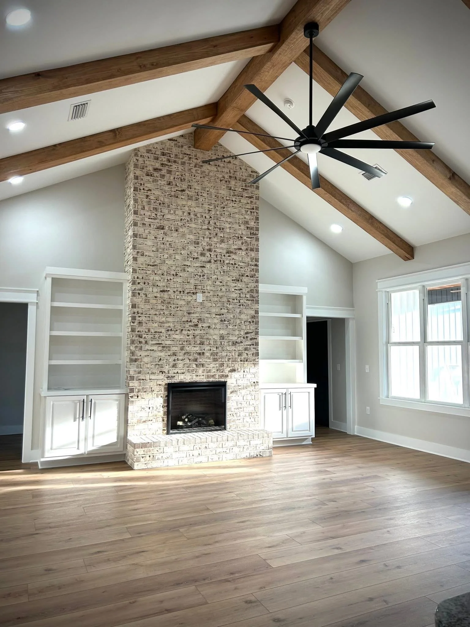 Interior of a living room with wooden vaulted ceiling, a modern black ceiling fan, a brick fireplace, white built-in shelves, and a large window letting in natural light.