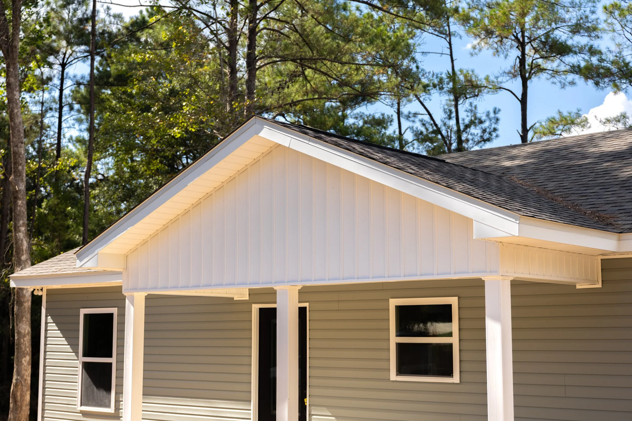 Front view of a house with beige siding, multiple windows, and a gable roof with asphalt shingles, surrounded by tall trees and blue sky.