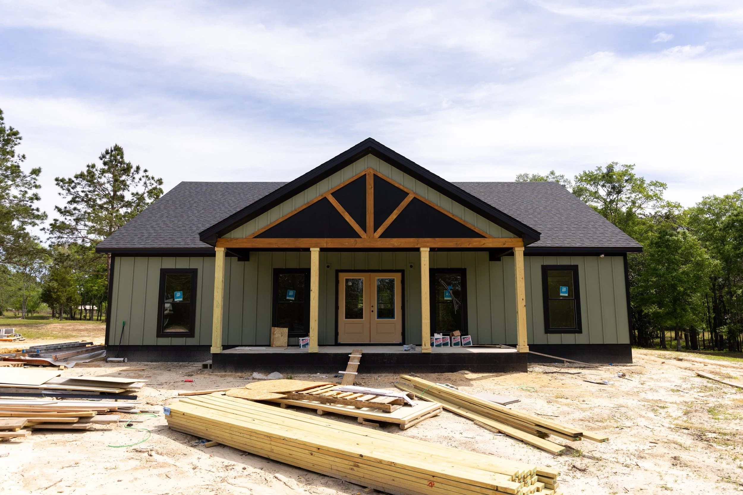 A house under construction with a front porch, showing unfinished wooden framing and building materials on the ground in front, trees in the background, and a partly cloudy sky.
