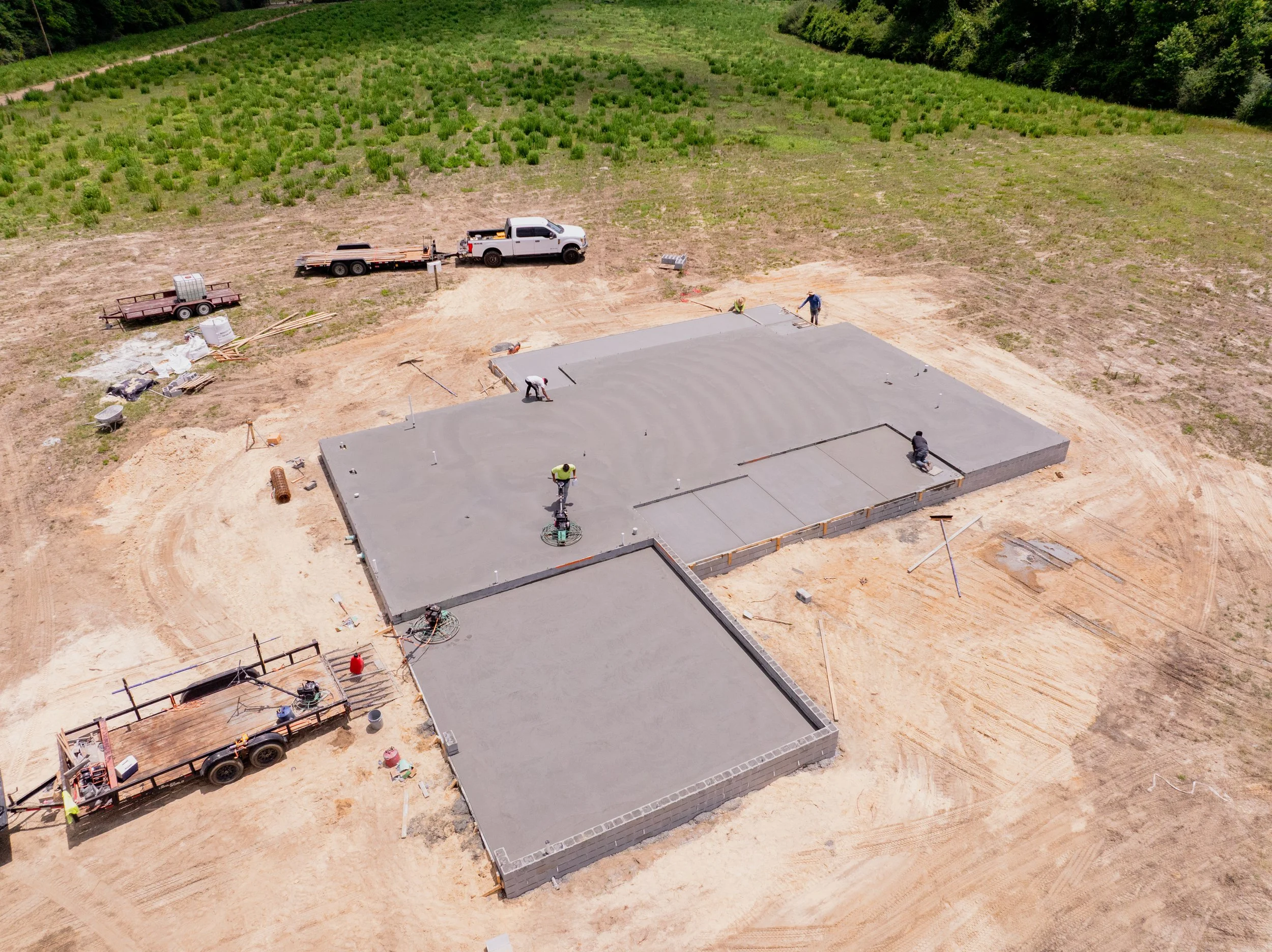 Aerial view of a construction site with a concrete foundation being poured, surrounded by dirt and grass, with workers and construction equipment present.