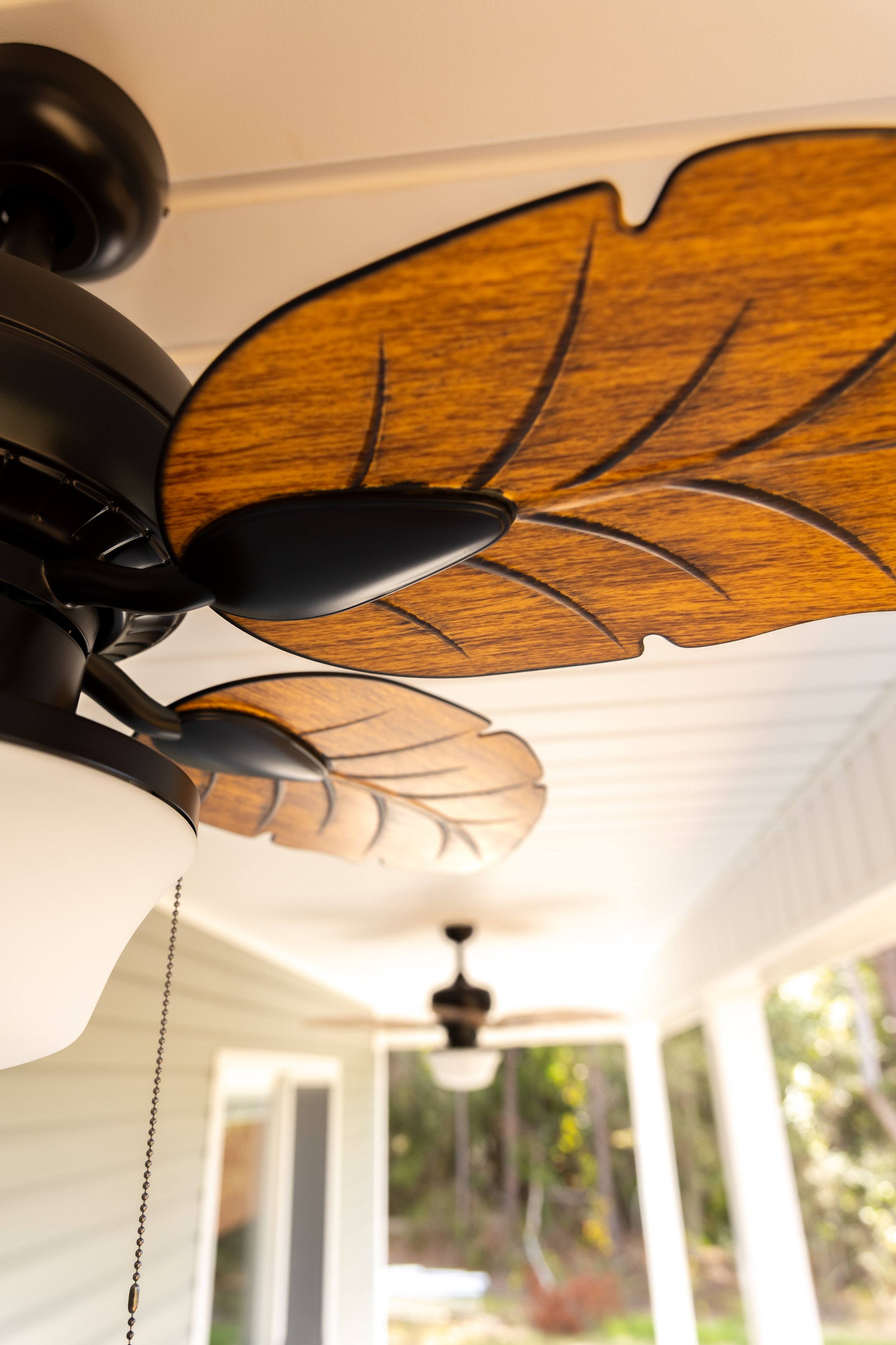 Close-up of a ceiling fan with leaf-shaped wooden blades, inside a house with visible windows and porch outside.