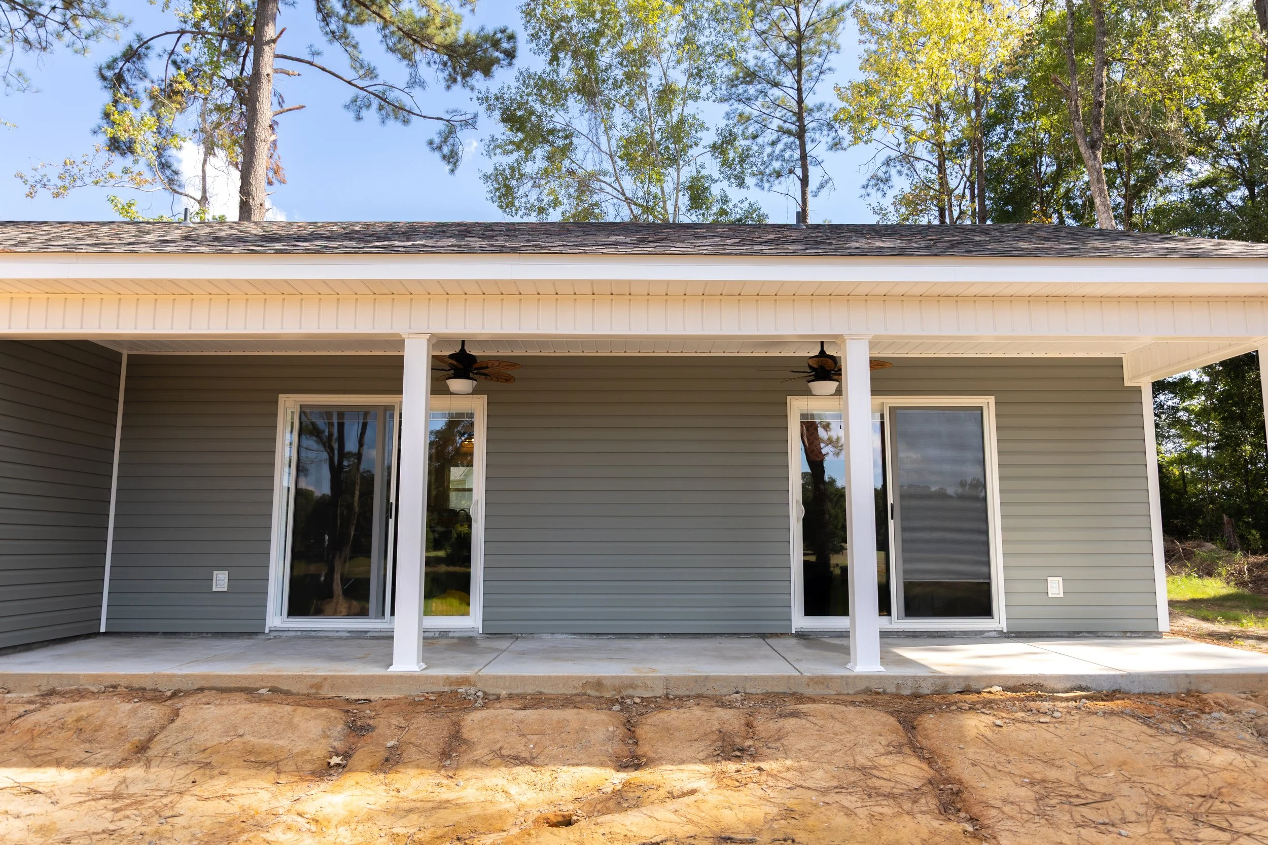 Back porch of a house with sliding glass doors, ceiling fans, and a concrete floor, with a wooded backyard visible through the doors.