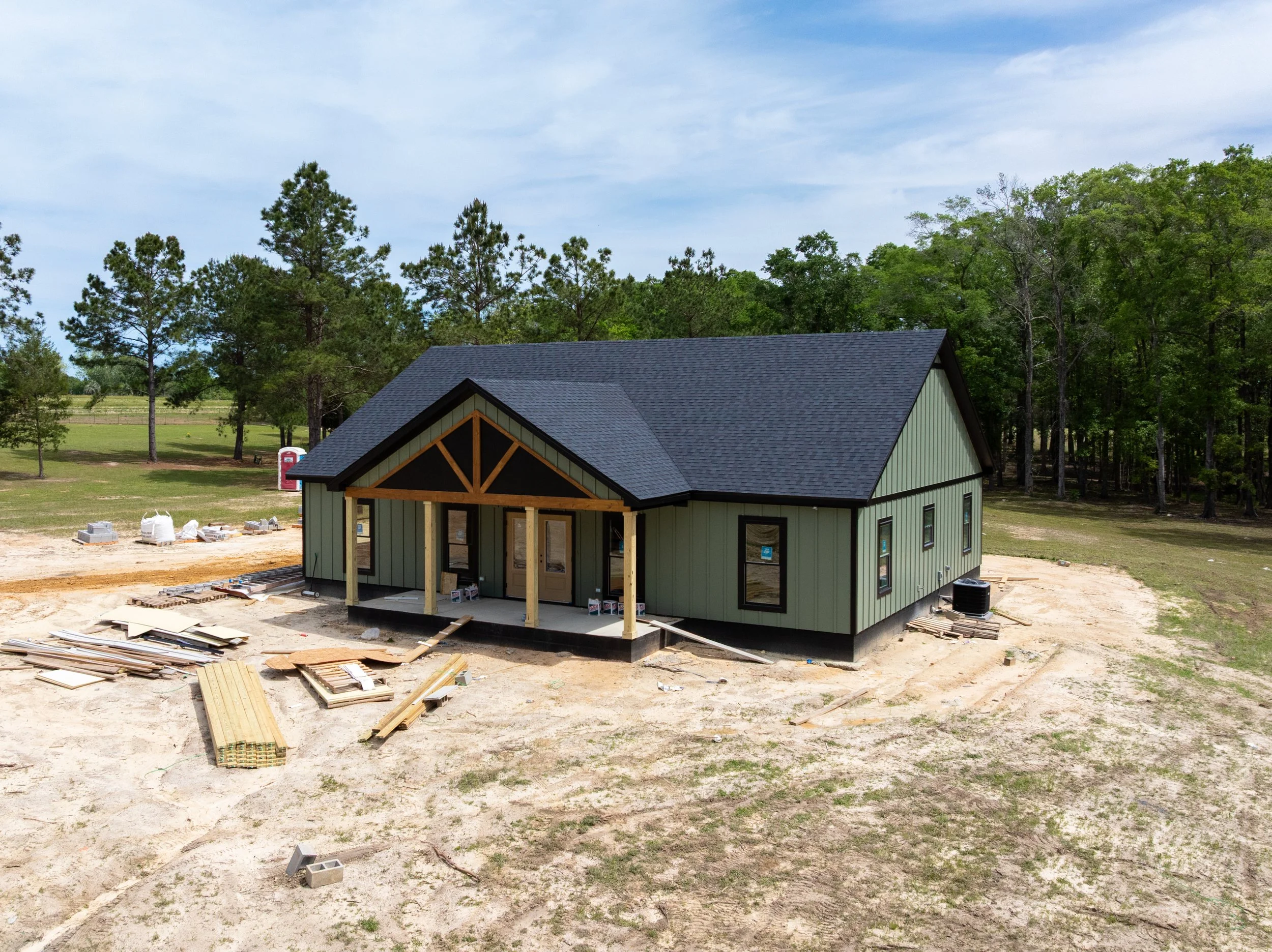 A house under construction with green siding, a black roof, and an open front porch in a grassy area surrounded by trees.