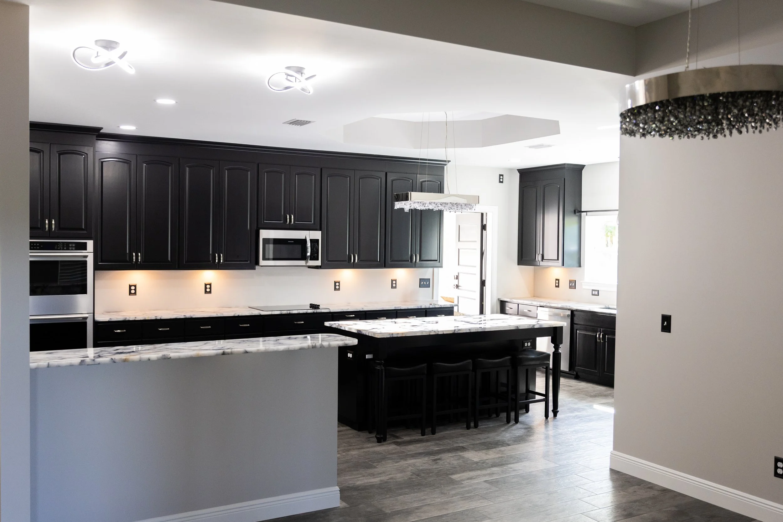 Modern kitchen with black cabinets, stainless steel appliances, white countertops with gray veining, and a marble countertop island with black stools. Recessed lighting and a unique hanging light fixture are visible.