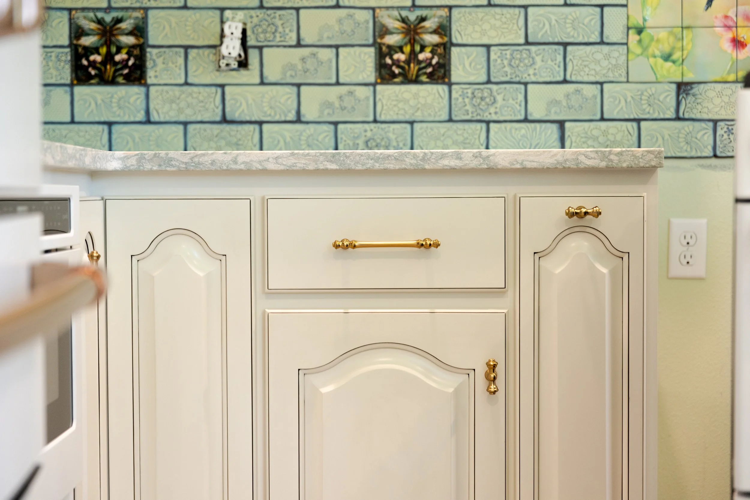 A kitchen countertop with white cabinets and gold hardware, a decorative tiled wall with floral and nature patterns in green and colorful accents.