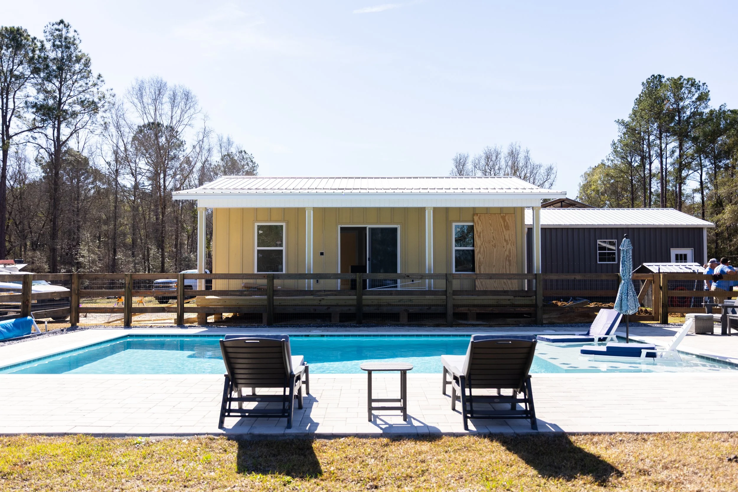 A backyard with a swimming pool, lounge chairs, an umbrella, and a fence. In the background, there are small houses or sheds, some under construction, with trees behind them.