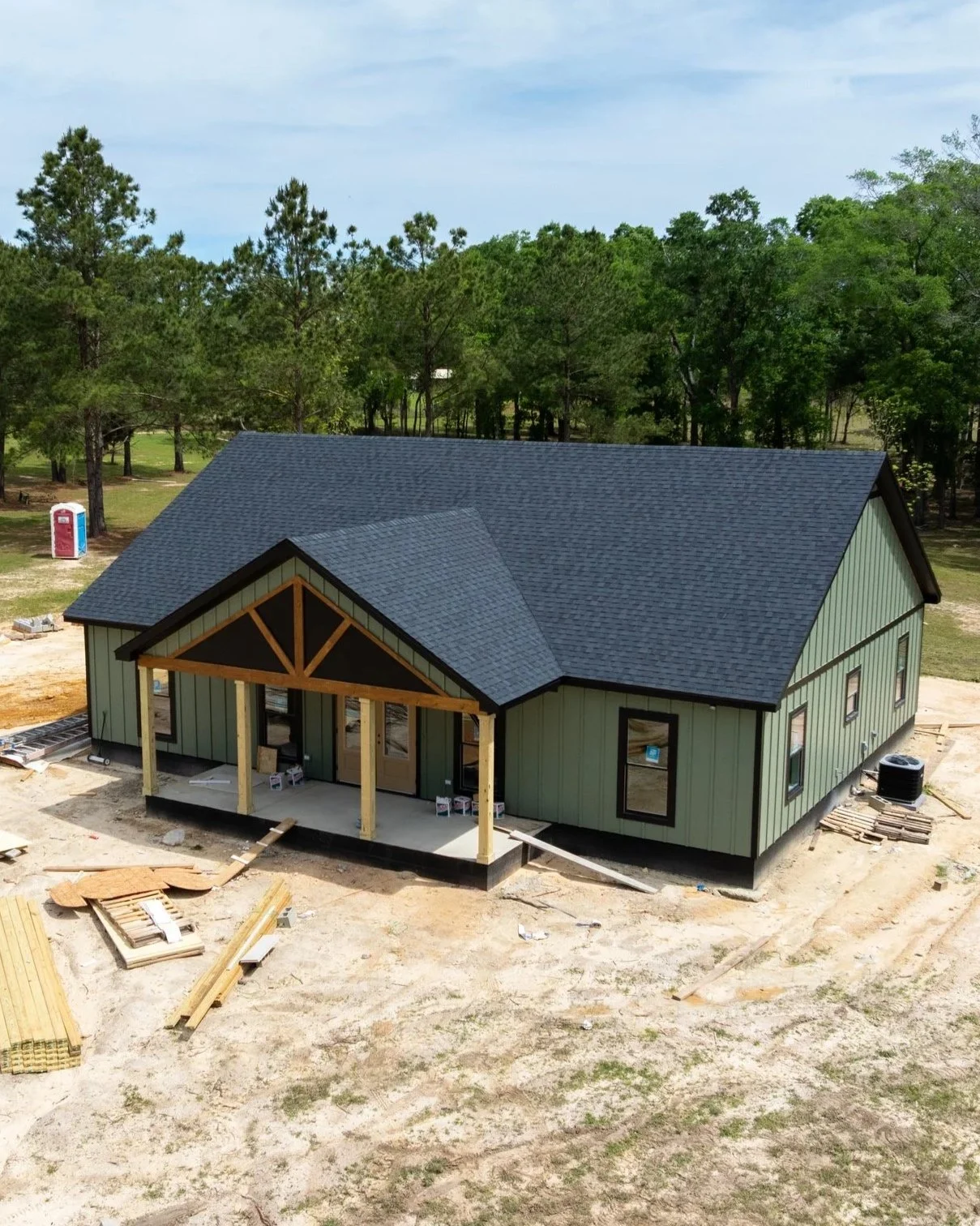 A house under construction with a black shingled roof and green siding, surrounded by a construction site with building materials, and a wooded area in the background.