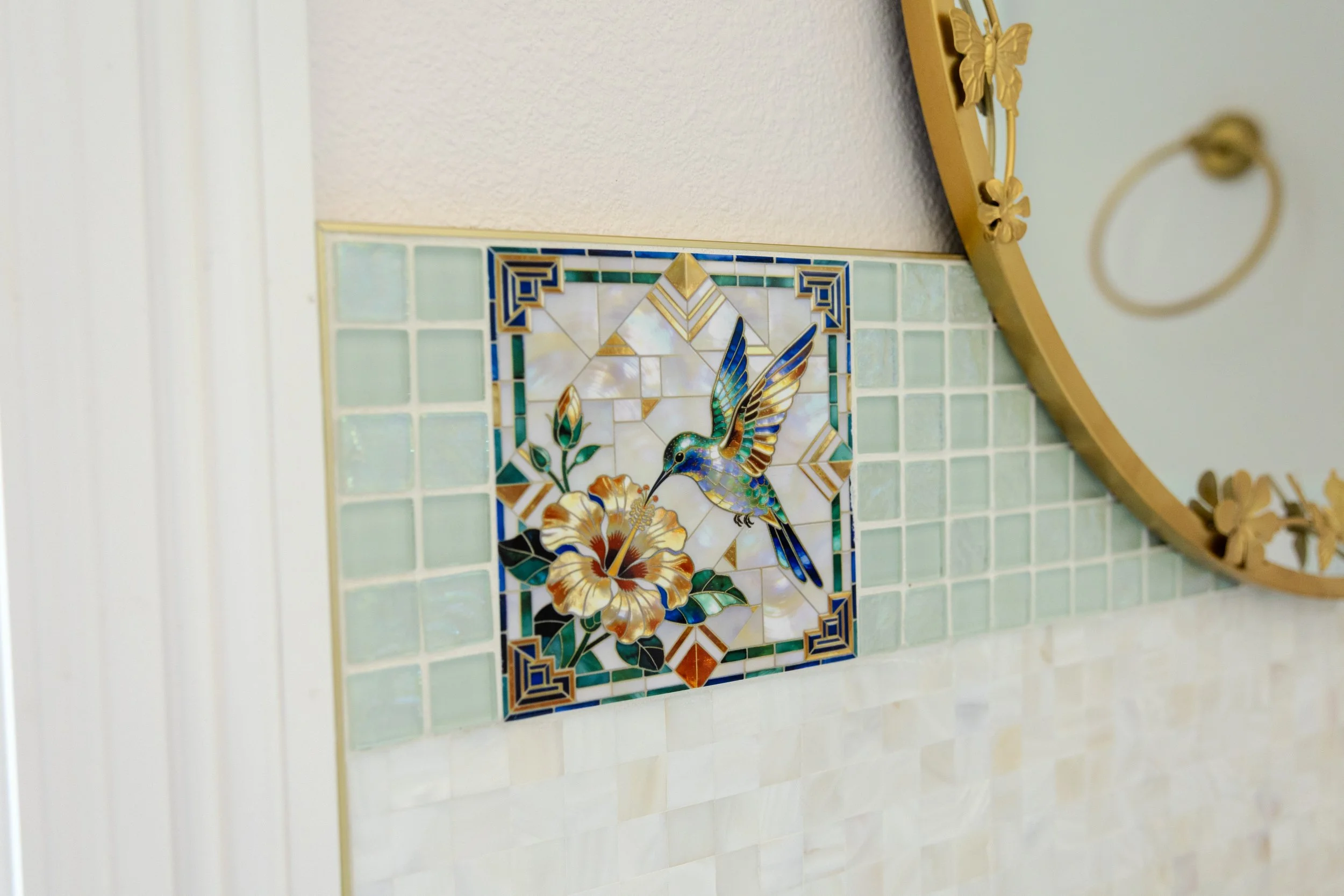 Close-up of a decorative mosaic tile featuring a hummingbird and flowers, surrounded by green and gold tiles, on a bathroom wall.