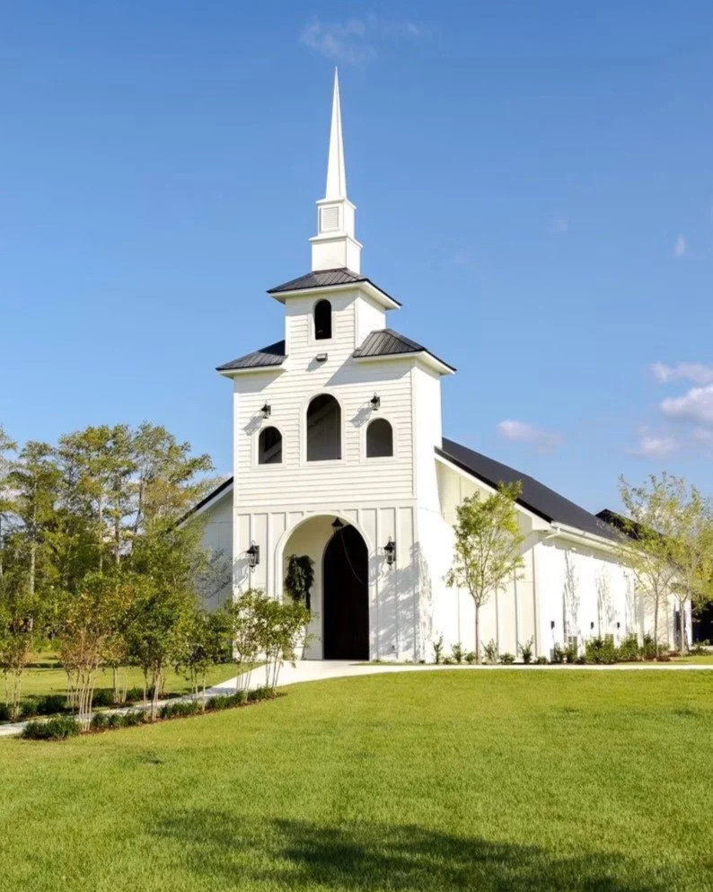White church with steeple, surrounded by green lawn and trees, under a blue sky.