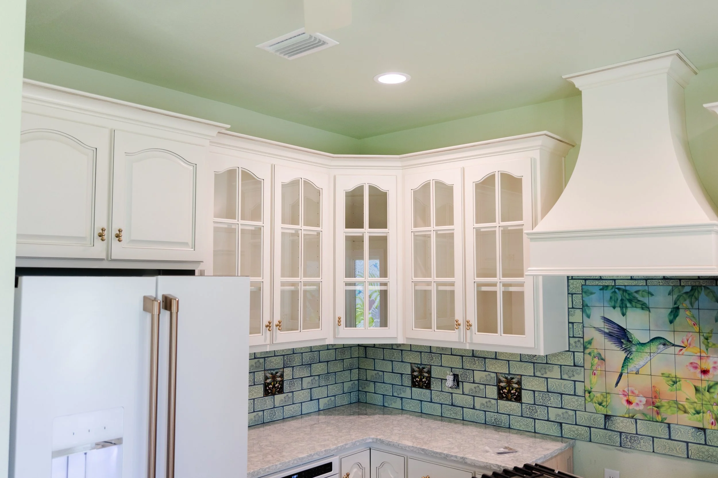 Kitchen with white cabinets and a colorful tile backsplash featuring birds and flowers, green walls, and a white range hood.