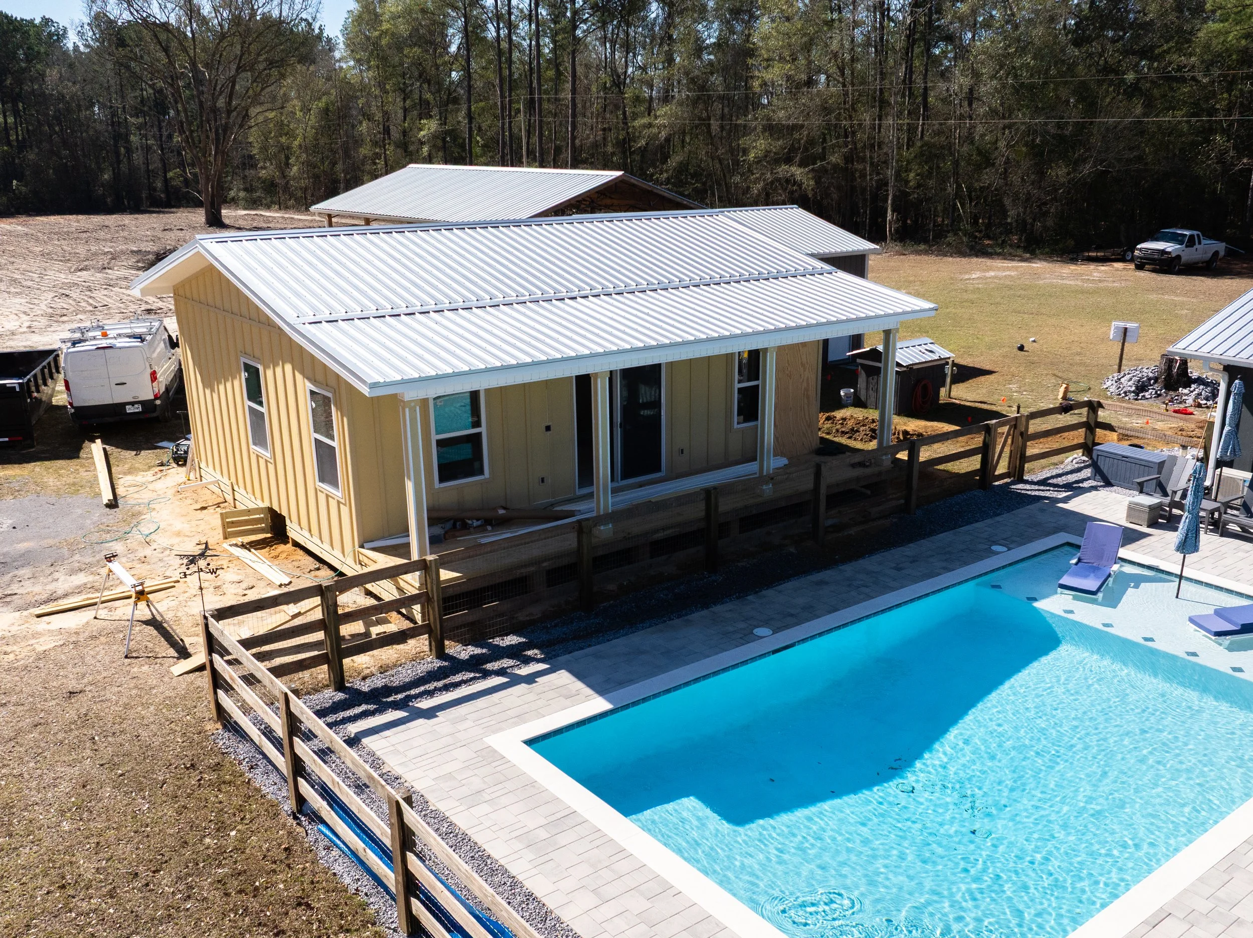 A backyard with a newly constructed yellow house that has a metal roof, a pool with a paved deck, a fenced yard, and a wooded background. Construction materials and vehicles are nearby.