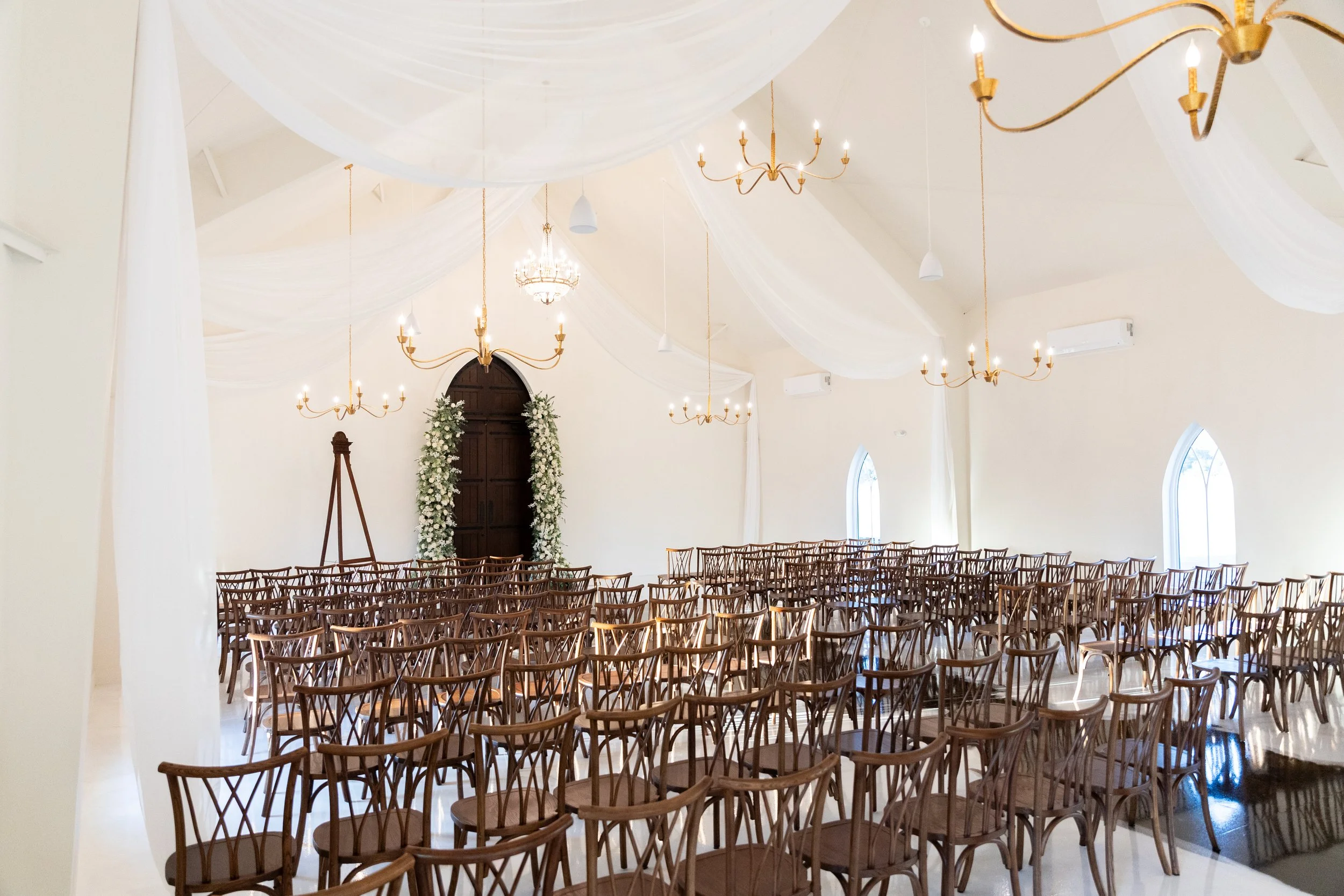 An elegant wedding venue with rows of wooden chairs facing an altar decorated with white flowers. The cream-colored walls are adorned with gold chandeliers and draped white fabric ceiling treatment. There are tall, arched windows on the side walls, a