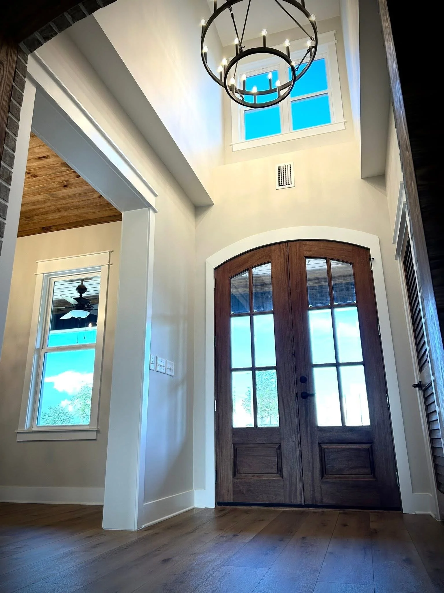 View of the interior entrance of a house with a wooden front door, windows on each side, a window above, a chandelier hanging from the ceiling, and wooden flooring.