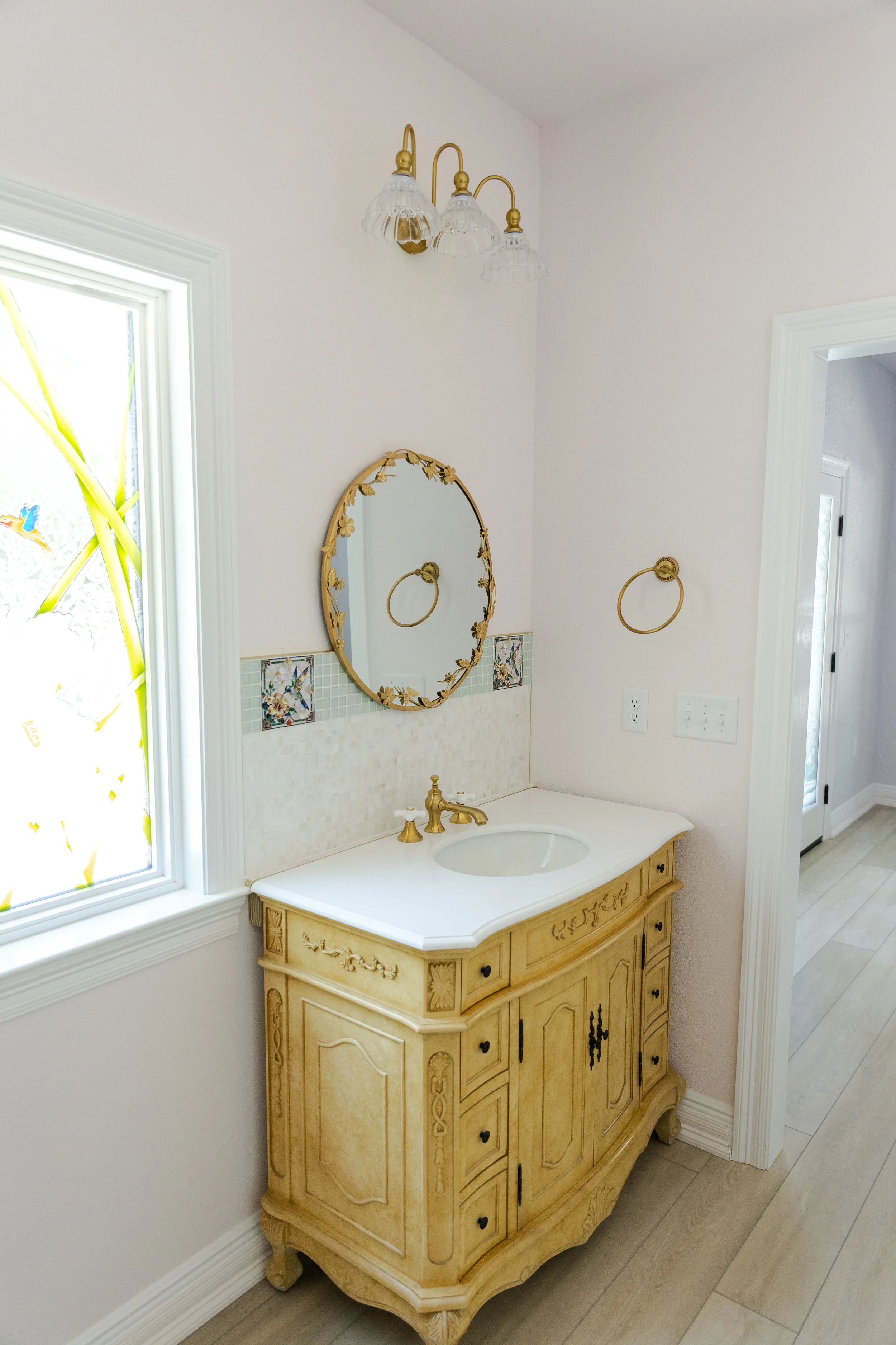 Decorative bathroom vanity with a mirror, gold accents, and a cream-colored wooden cabinet, alongside a stained glass window with floral and bird design.