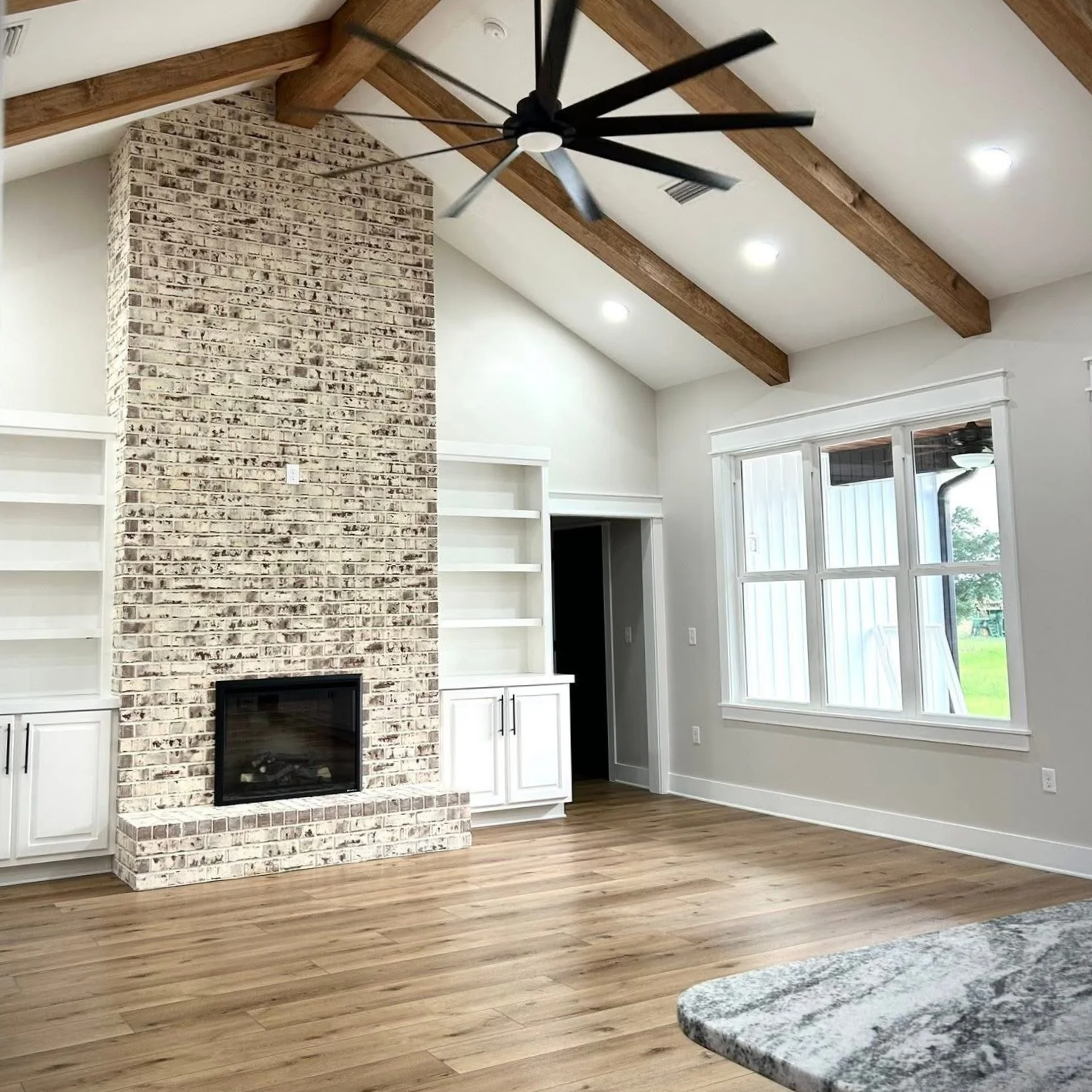 Interior living room with a brick fireplace, built-in white shelves and cabinets, large window, wooden ceiling beams, and hardwood floors.