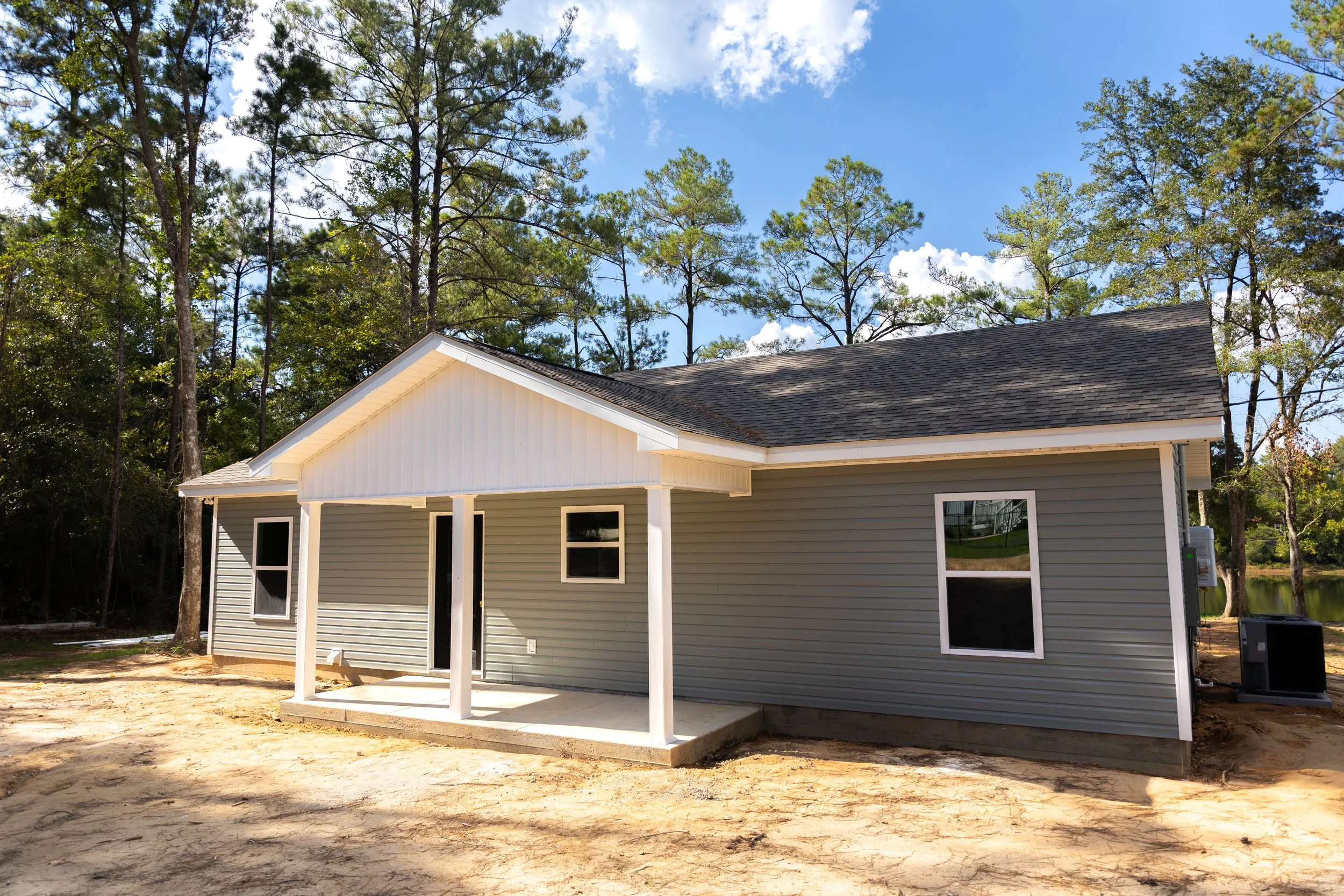 A house under construction with grey siding and a dark shingled roof, surrounded by trees and a clear blue sky.
