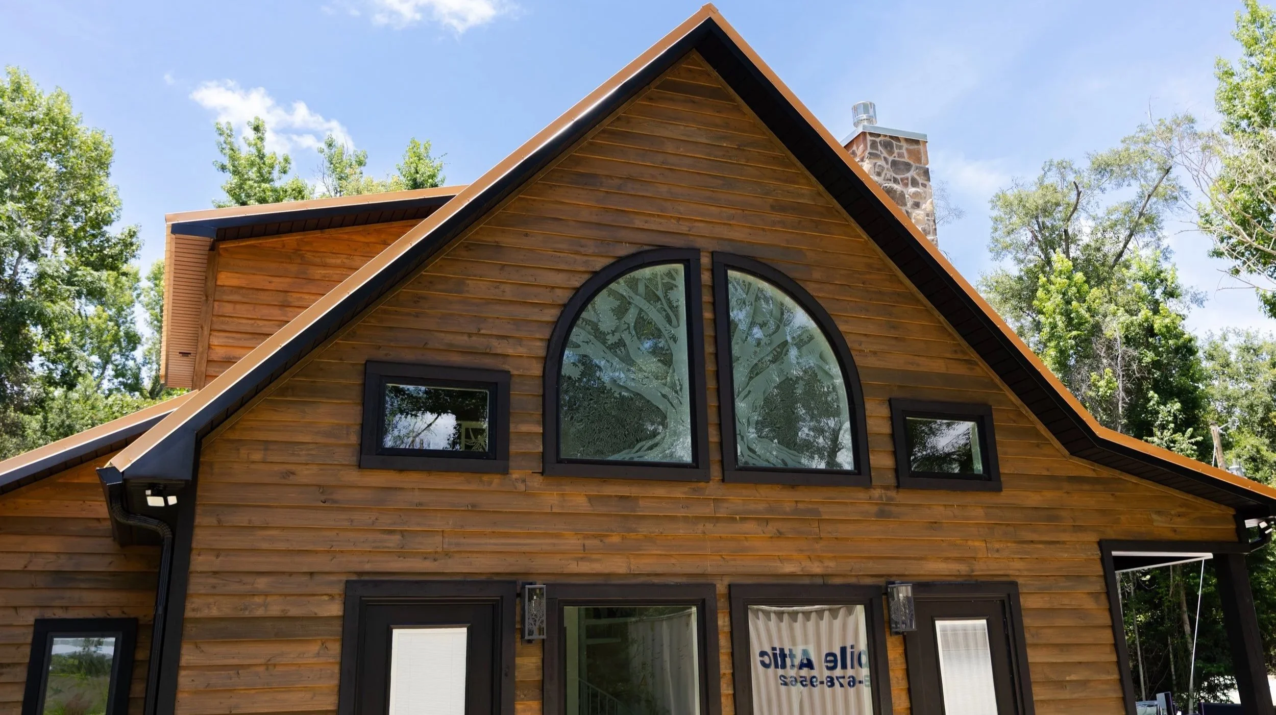 A wooden house with a steep gable roof, large arched window, and surrounding trees under a blue sky.