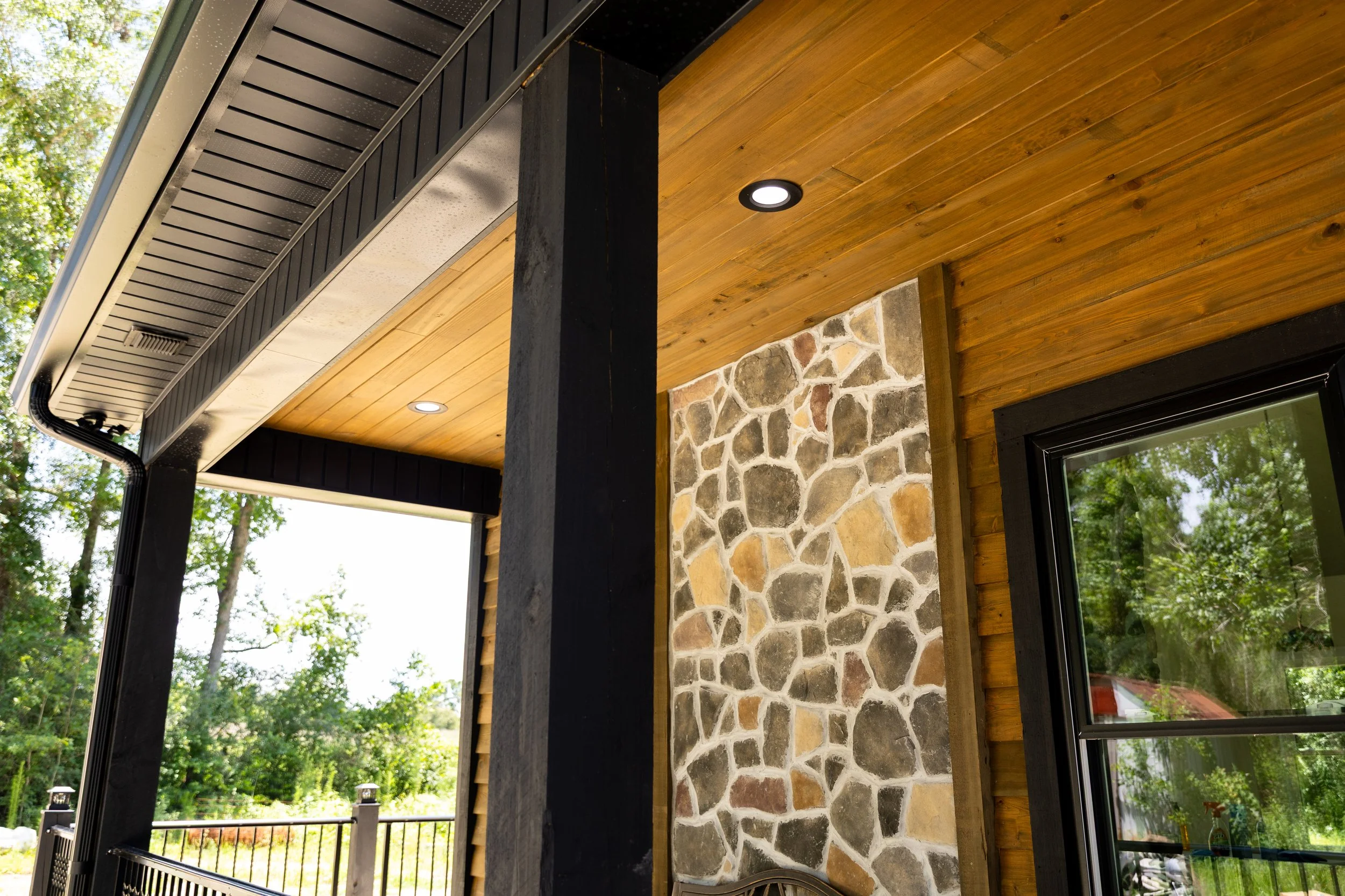 Close-up view of the exterior porch of a house featuring a wooden ceiling, a brick and stone wall, and a window. The porch has recessed lighting and black structural supports.