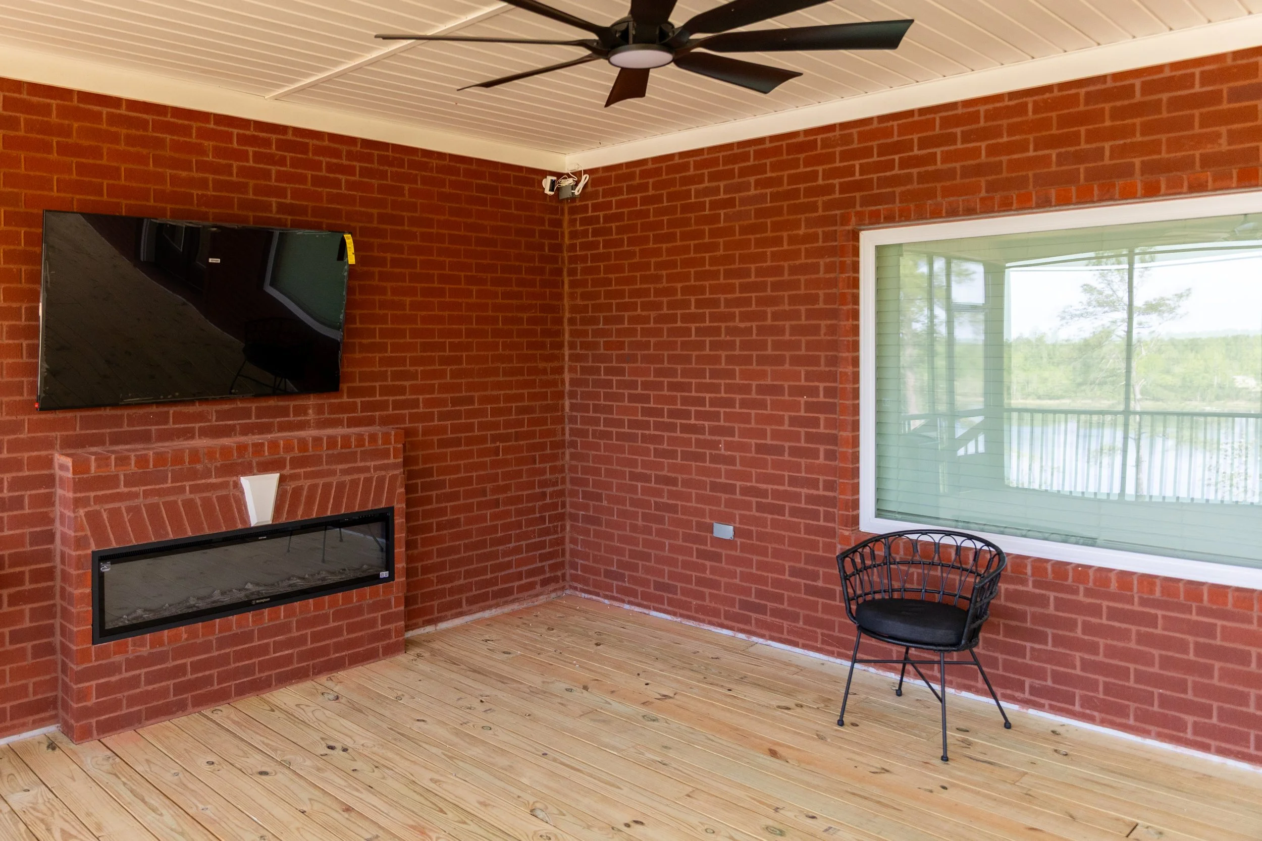 A corner of a room with red brick walls, a wooden floor, and a white ceiling with a black ceiling fan. There is a mounted flat-screen TV and a fireplace beneath it. A large window with green blinds overlooks trees outside, and a black chair is positi
