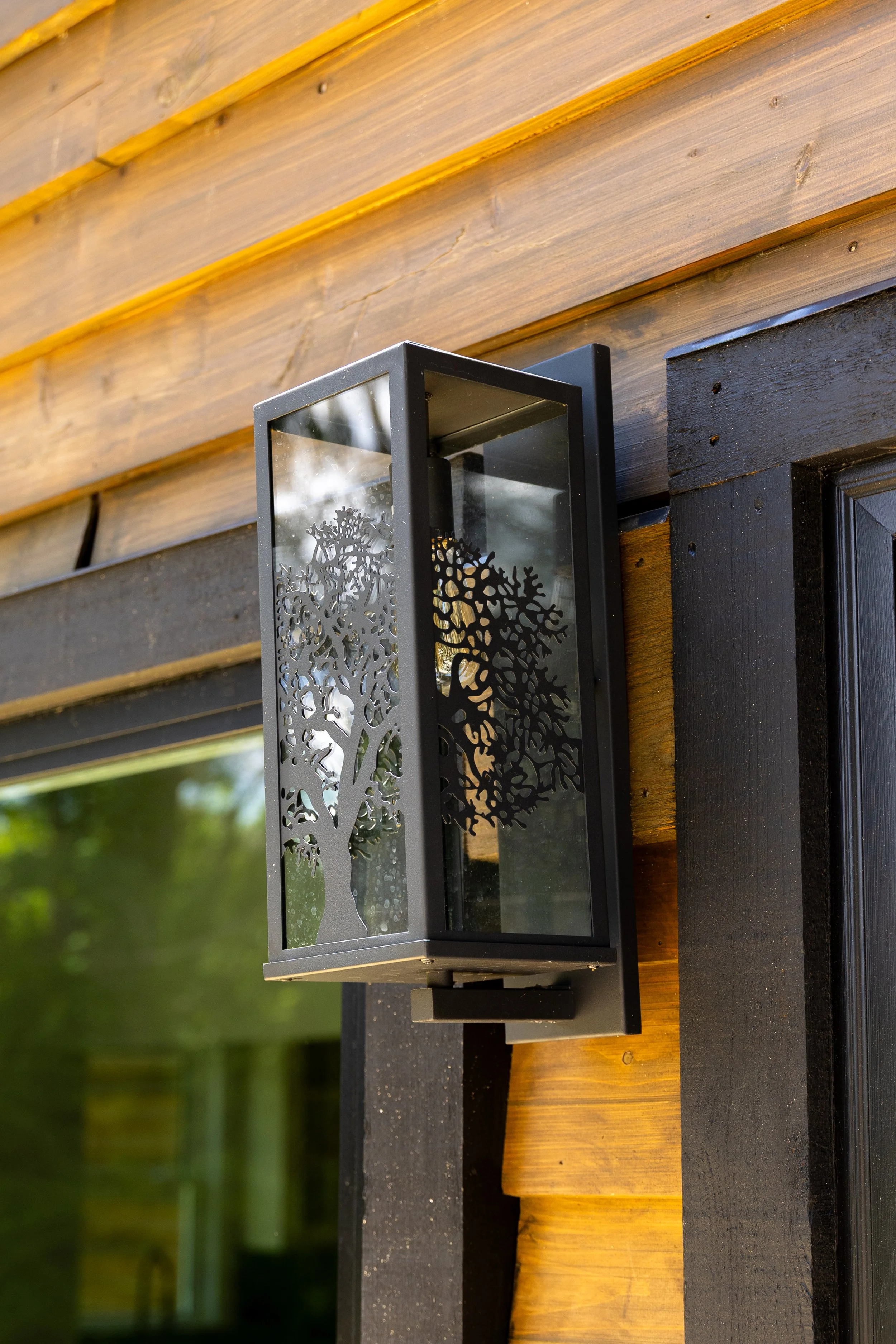 Close-up of a modern decorative black metal wall-mounted lantern with a cut-out tree design, mounted on wood and black painted wall, outdoors during daytime.