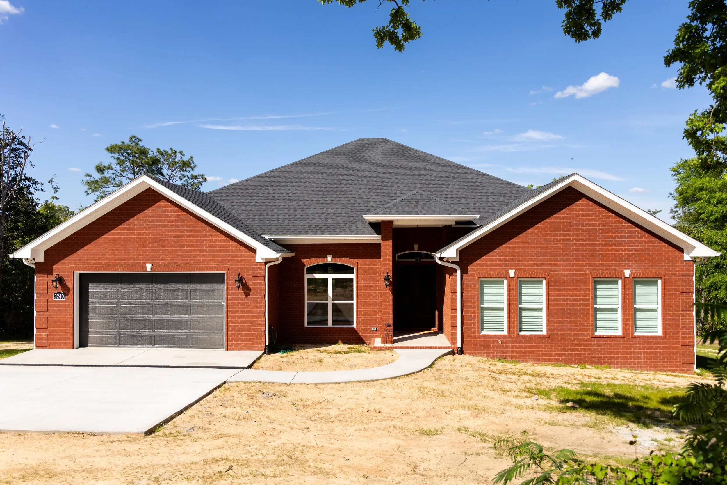 Front view of a two-story red brick house with a dark gray shingled roof, metallic garage door, black front door, and several windows with blinds, under a blue sky with a few clouds, surrounded by green trees and a front yard with bare dirt.
