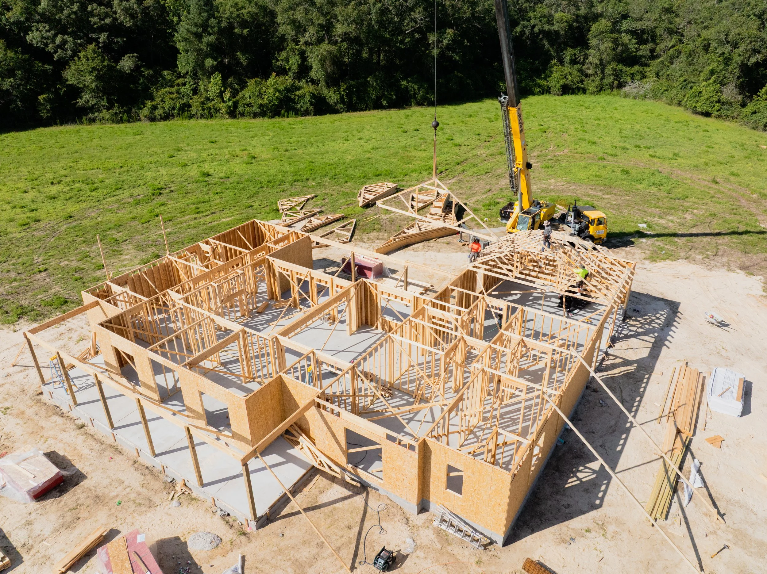 Aerial view of a house under construction with wooden framing and construction workers on site, surrounded by green fields and trees.