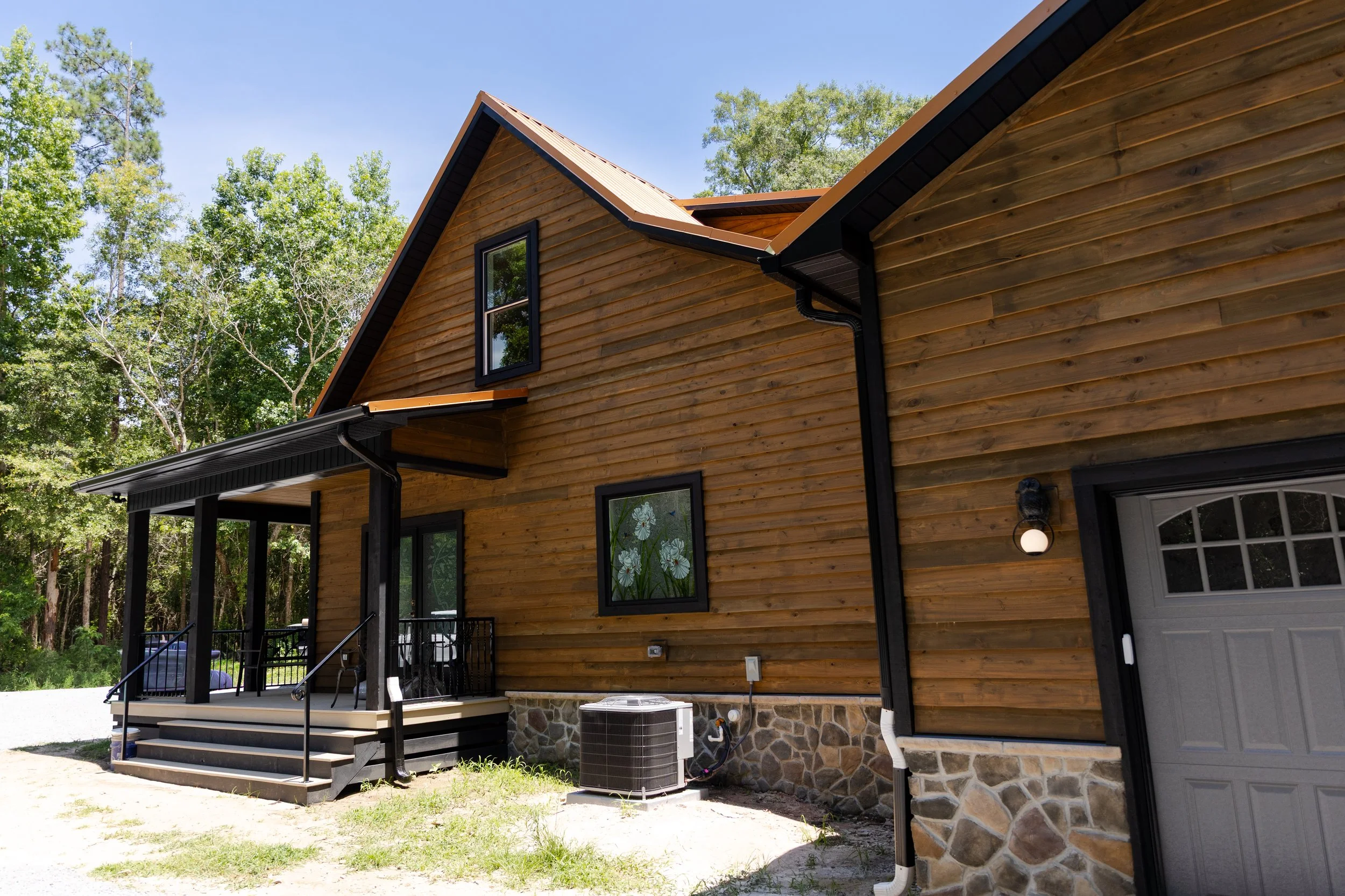 The backside of a wooden house with a small porch, black window frames, and a stone foundation. The house has a sloped roof and is surrounded by trees under a clear blue sky.