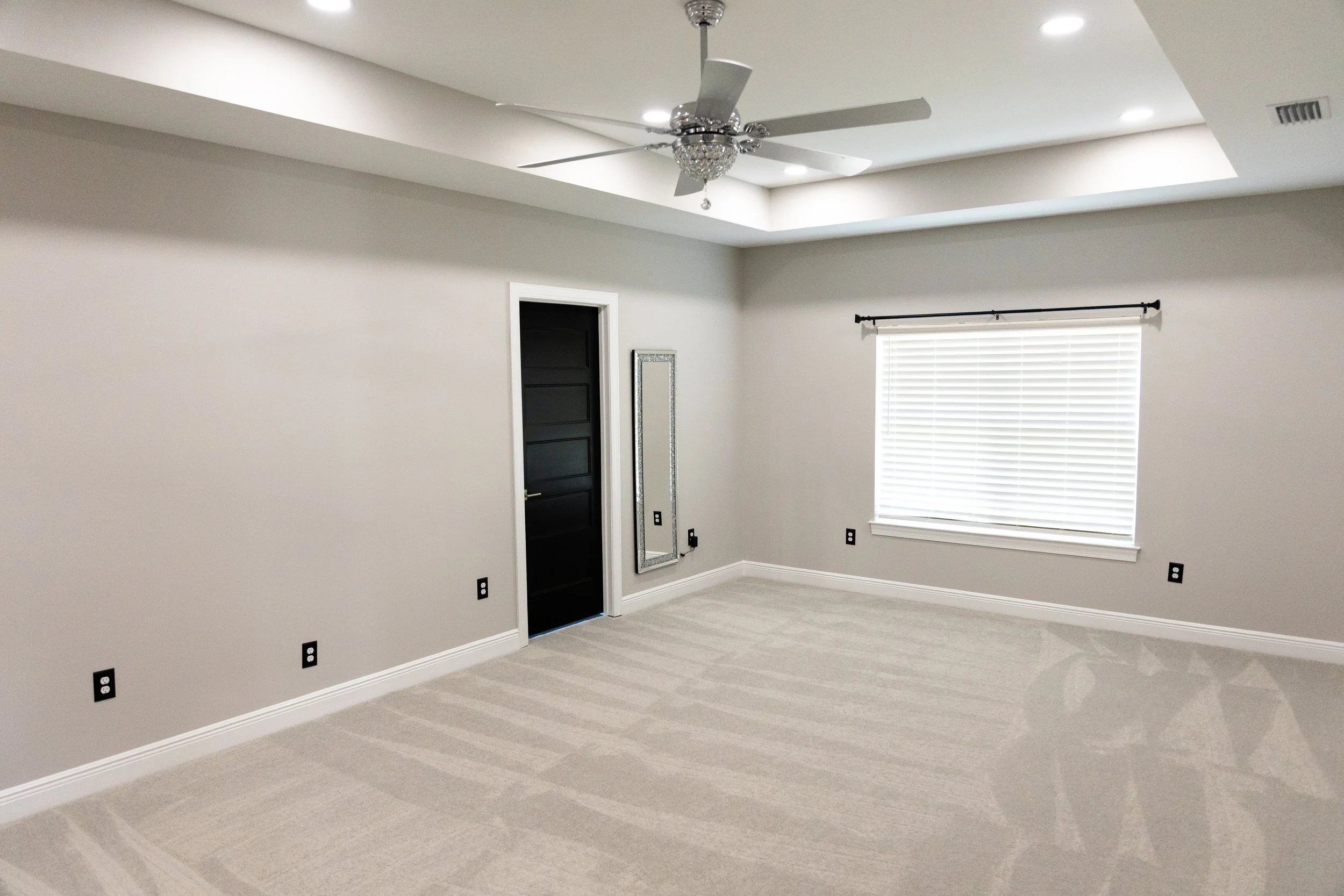 Empty room with beige walls, a large window with blinds, a ceiling fan, and multiple electrical outlets.